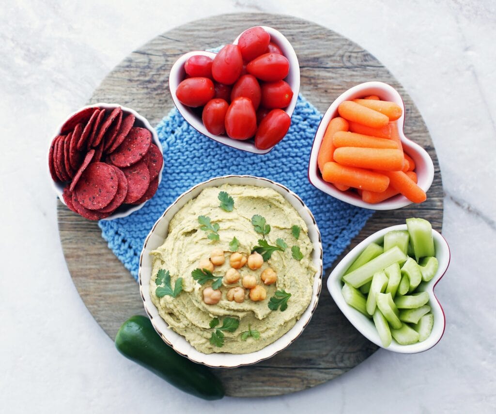 An overhead shot of a bowl of jalapeno cilantro hummus with carrots, tomatoes, celery, and crackers.