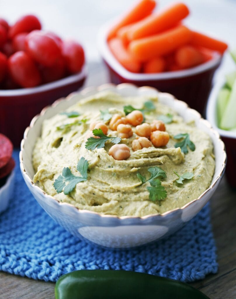 A closeup view of a bowl of jalapeno cilantro hummus with vegetables in the background.