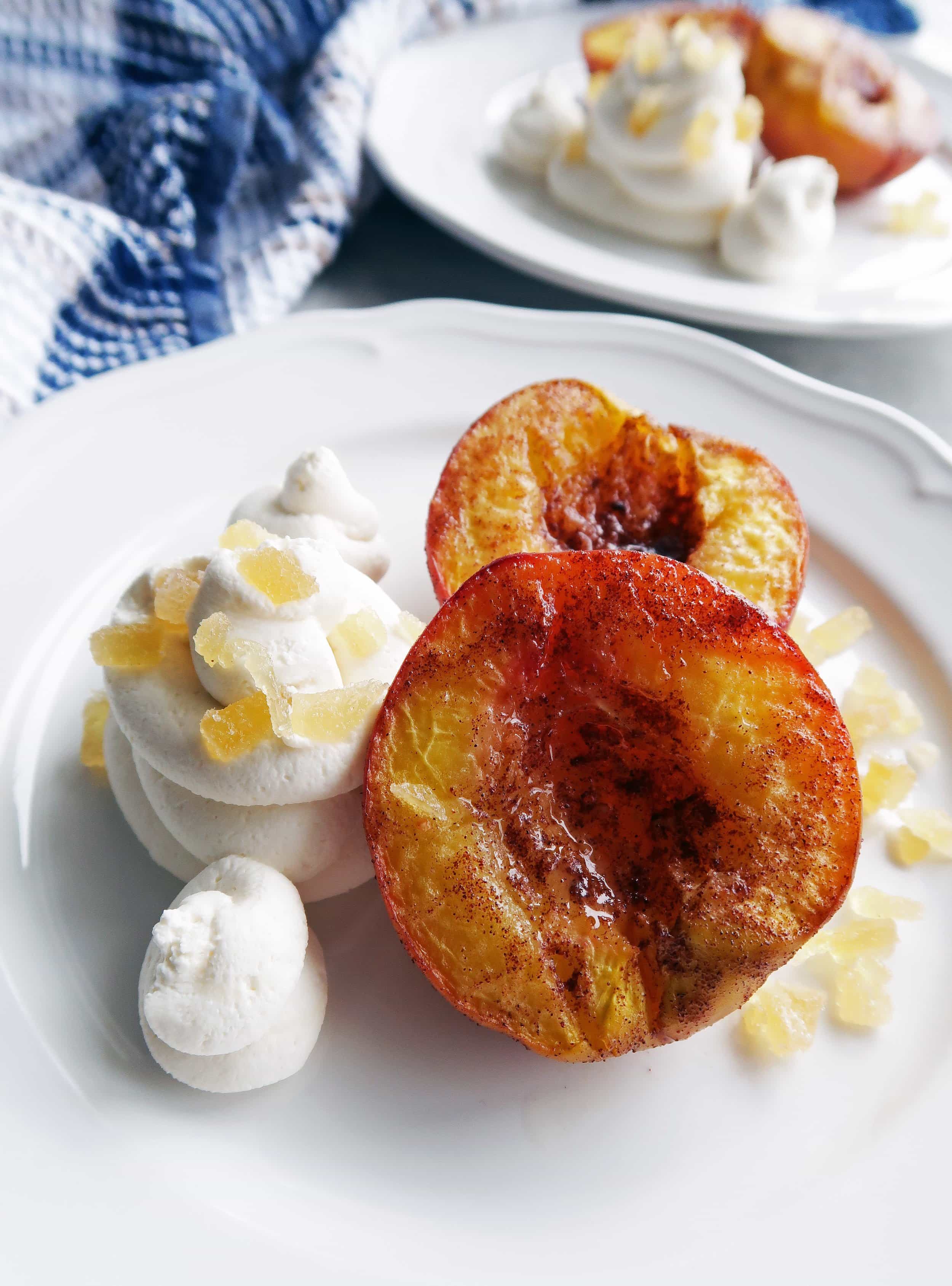 Closeup side view of Cinnamon Roasted Peach Halves with Honey Mascarpone Whipped Cream and crystallized ginger on a white plate with another similar plate in the background.