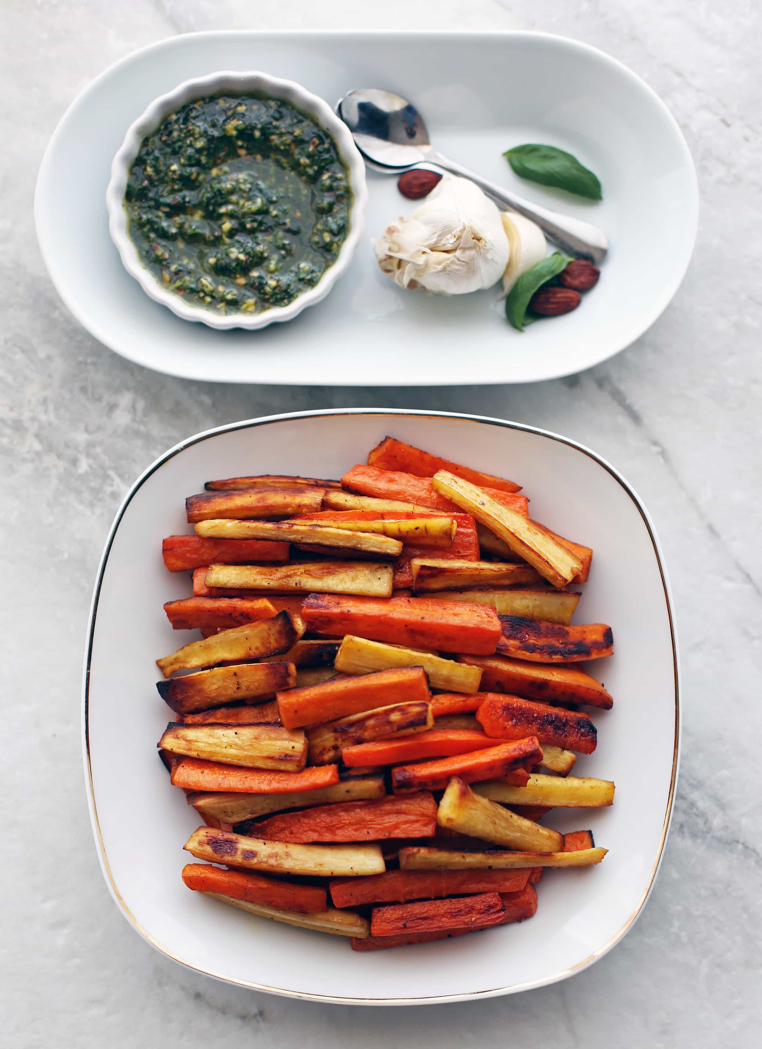 Roasted Balsamic Root Vegetables stacked on a white plate with basil almond pesto in a small bowl with spoons to its side.