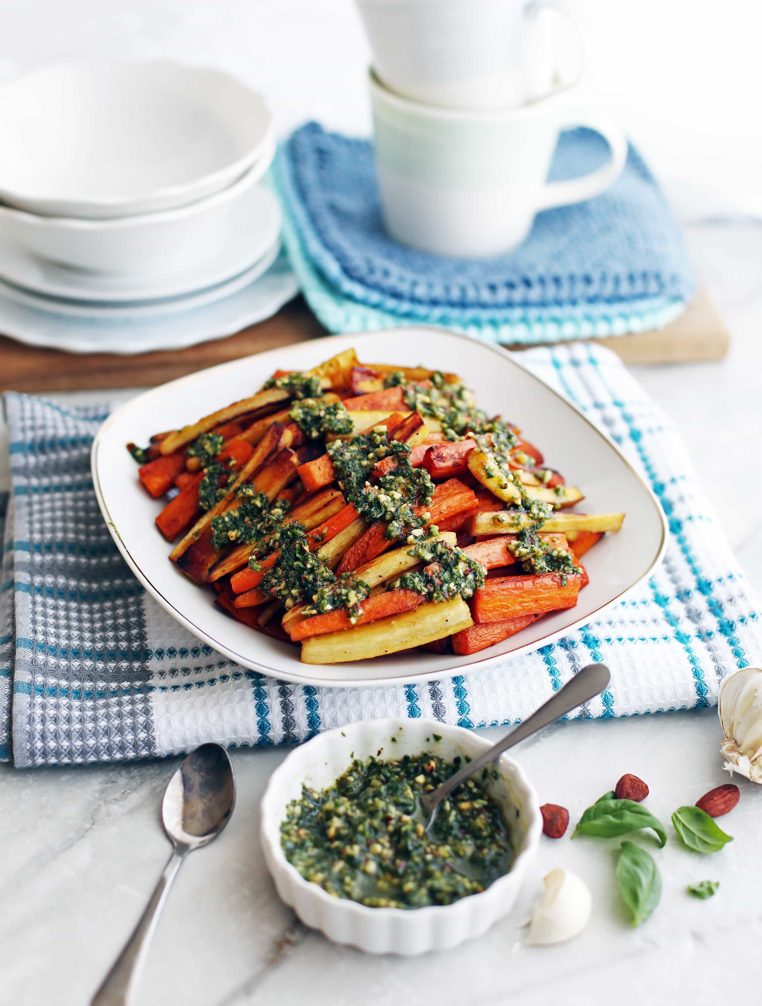 Roasted Balsamic Root Vegetables topped with Basil Almond Pesto on a white plate; more pesto in a small bowl on the side.