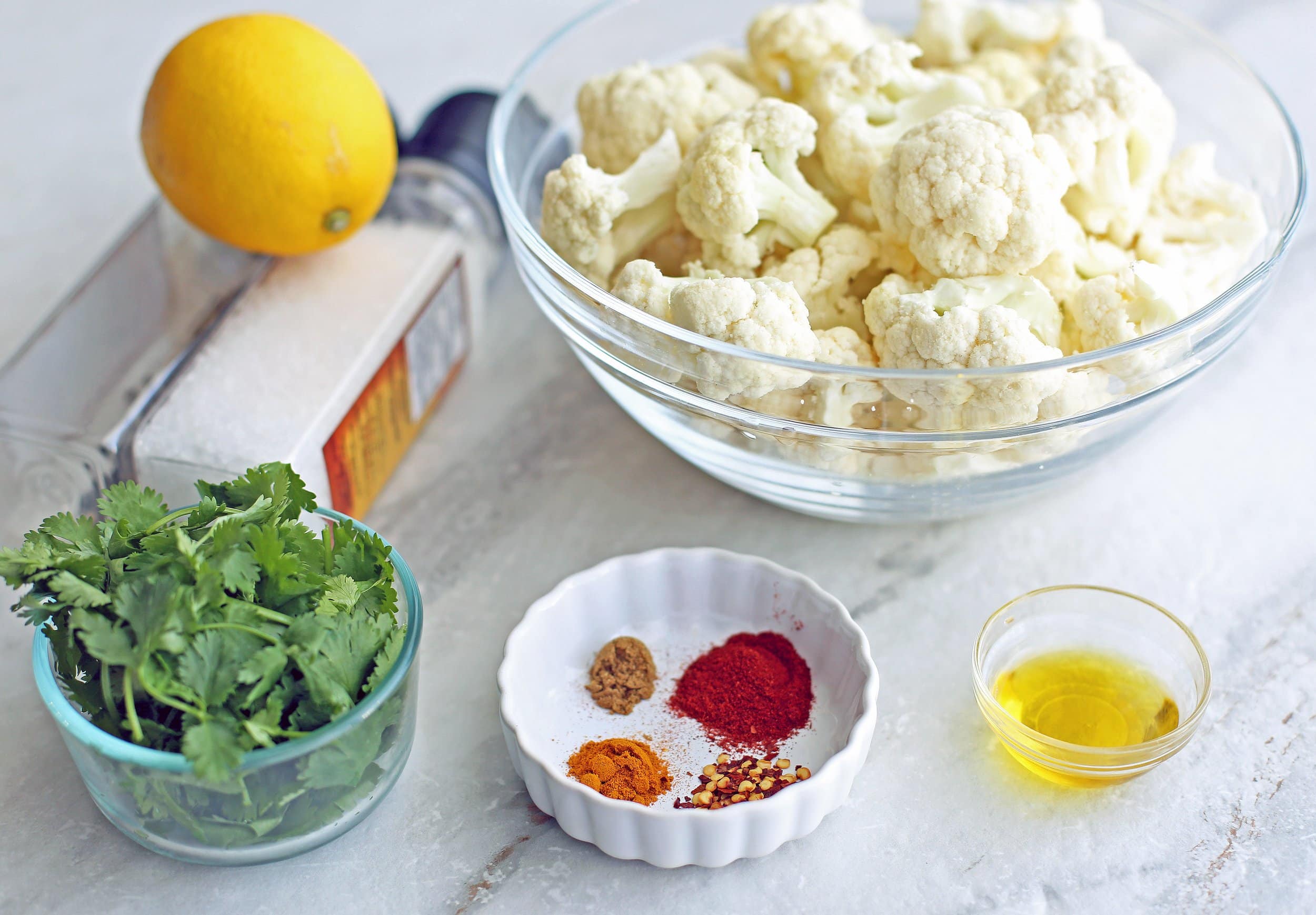 Side angled view of cauliflower florets, spices, lemon, olive oil, and cilantro.