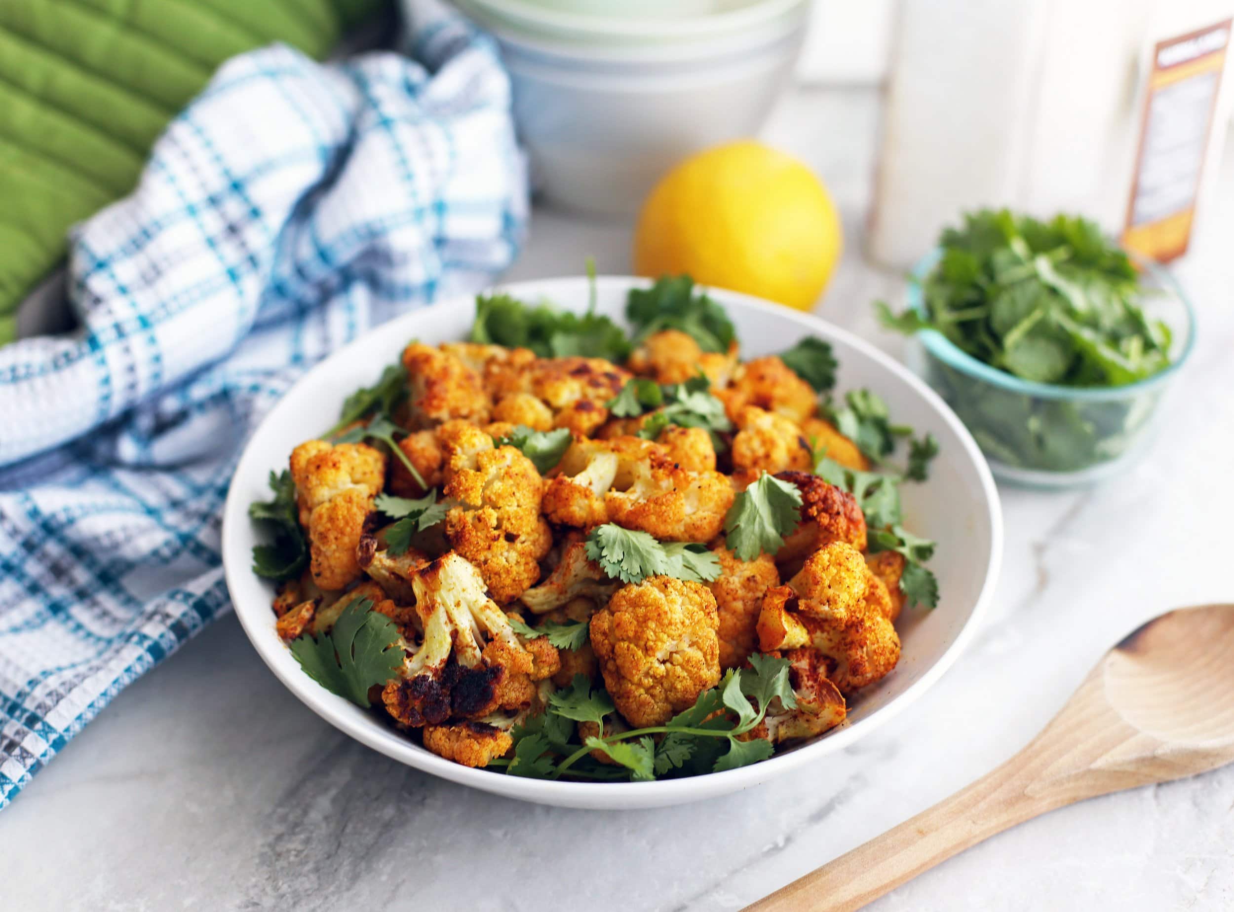 A wide bowl full of roasted spicy cauliflower with fresh cilantro with more cilantro and a lemon in the background.