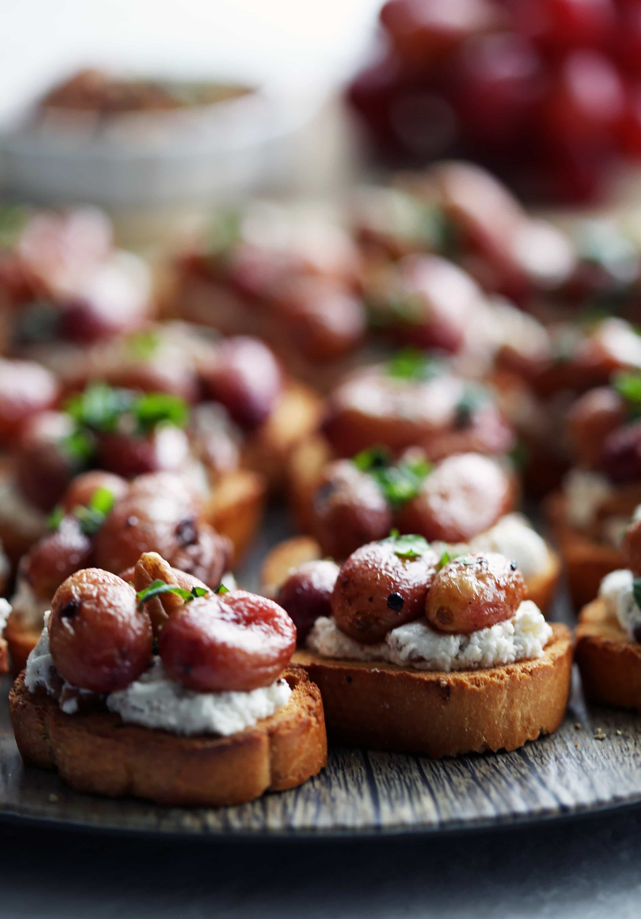 Side close-up of two roasted grape and goat cheese crostini with more crostini in the background.