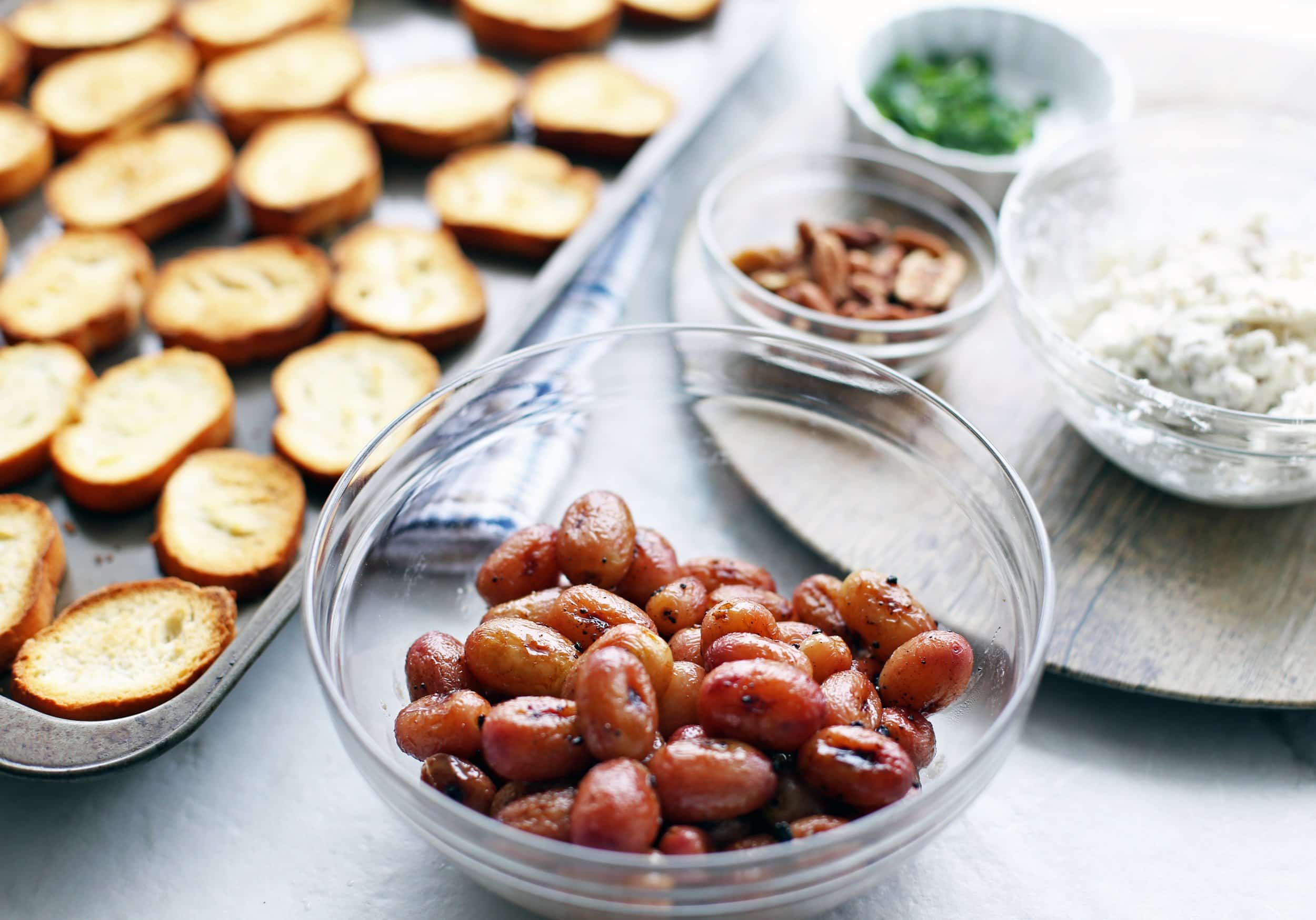 Balsamic roasted grapes in a glass bowl, toasted sliced baguette slices on a baking sheet, and goat cheese, pecans, and mint on a round platter.