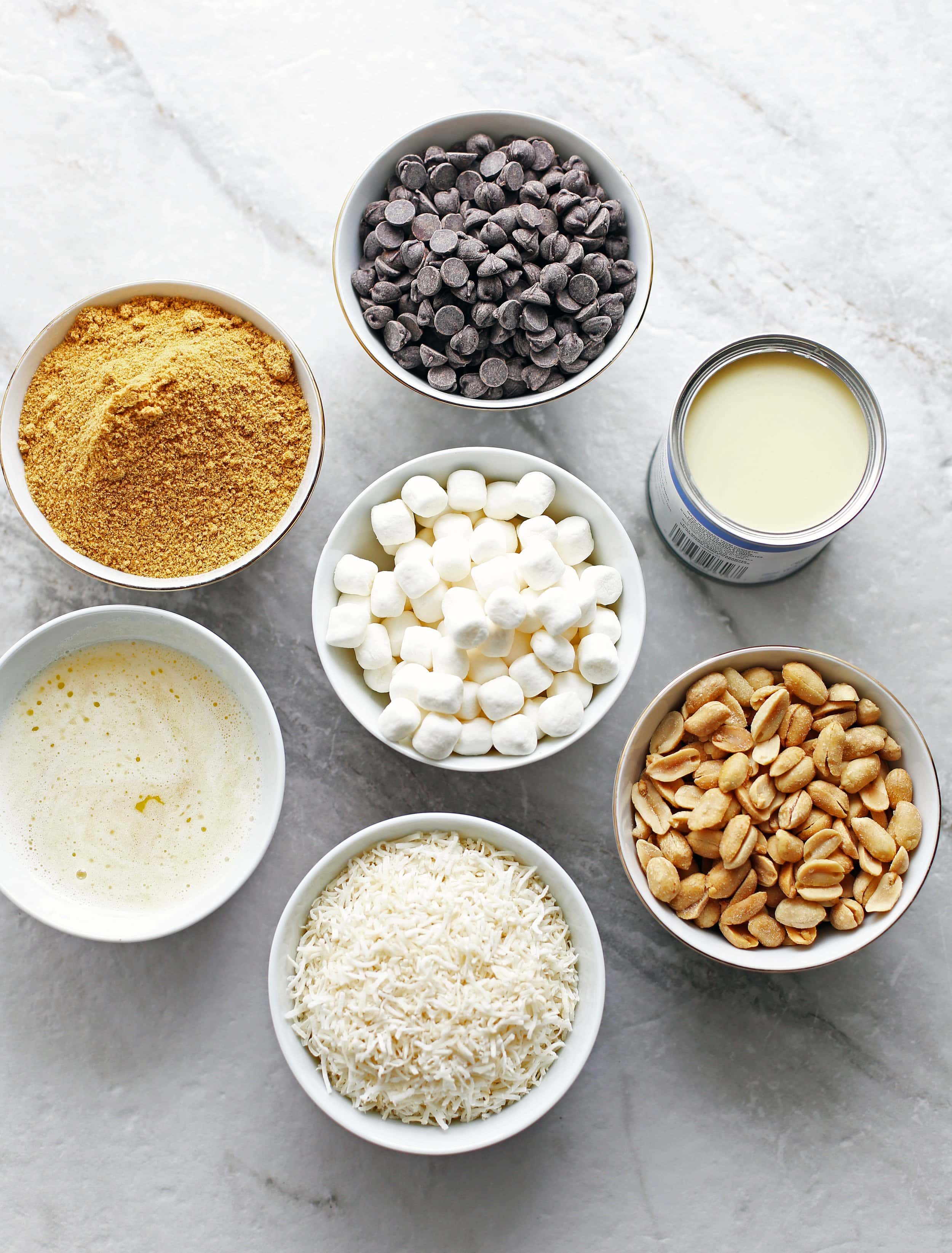 Bowls containing chocolate chips, peanuts, shredded coconut, marshmallows, condensed milk, graham cracker crumbs, and melted butter.