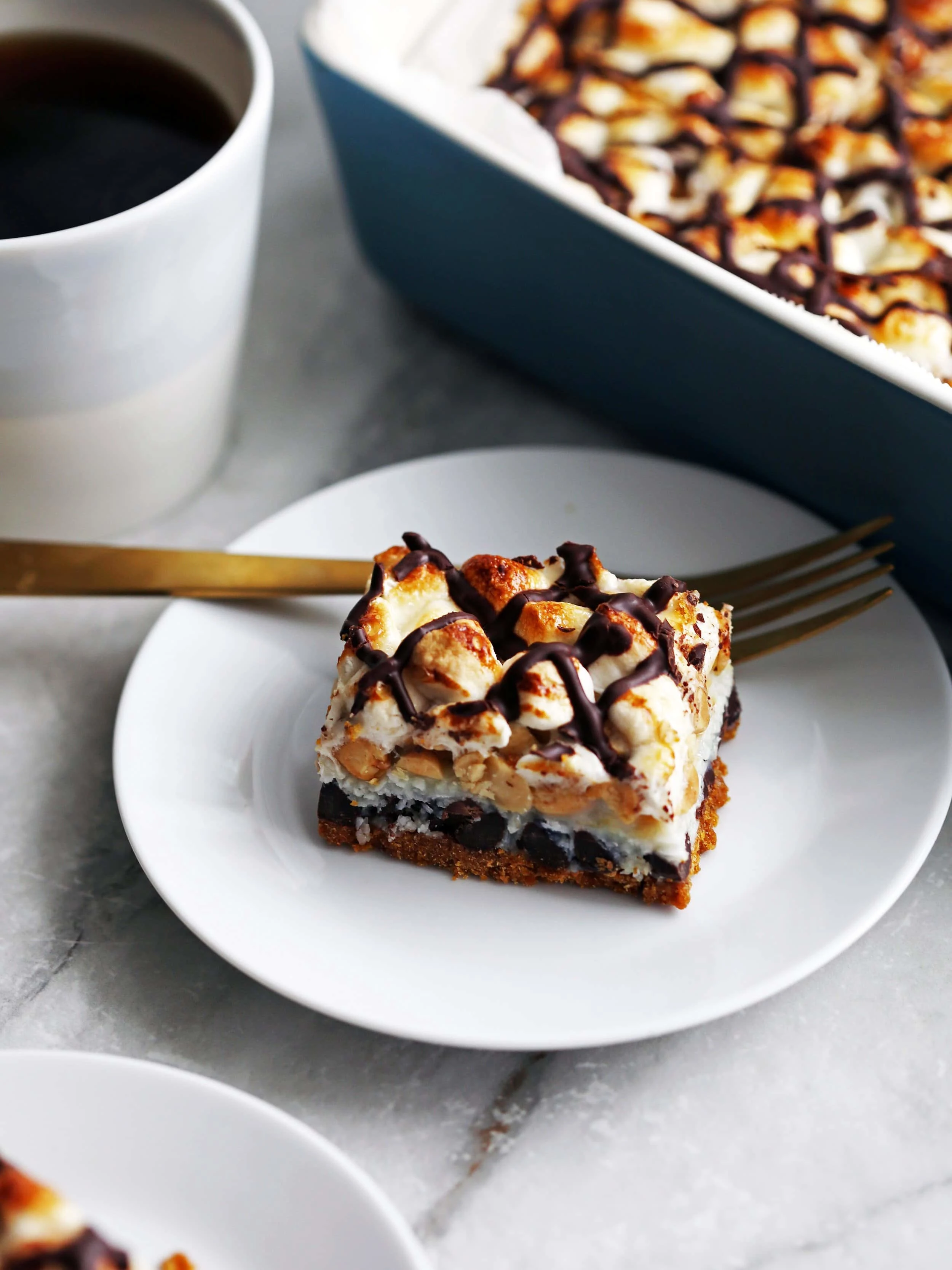 A side angled view of a seven layer rocky road magic bar on a white plate with a fork with more magic bars in the background.