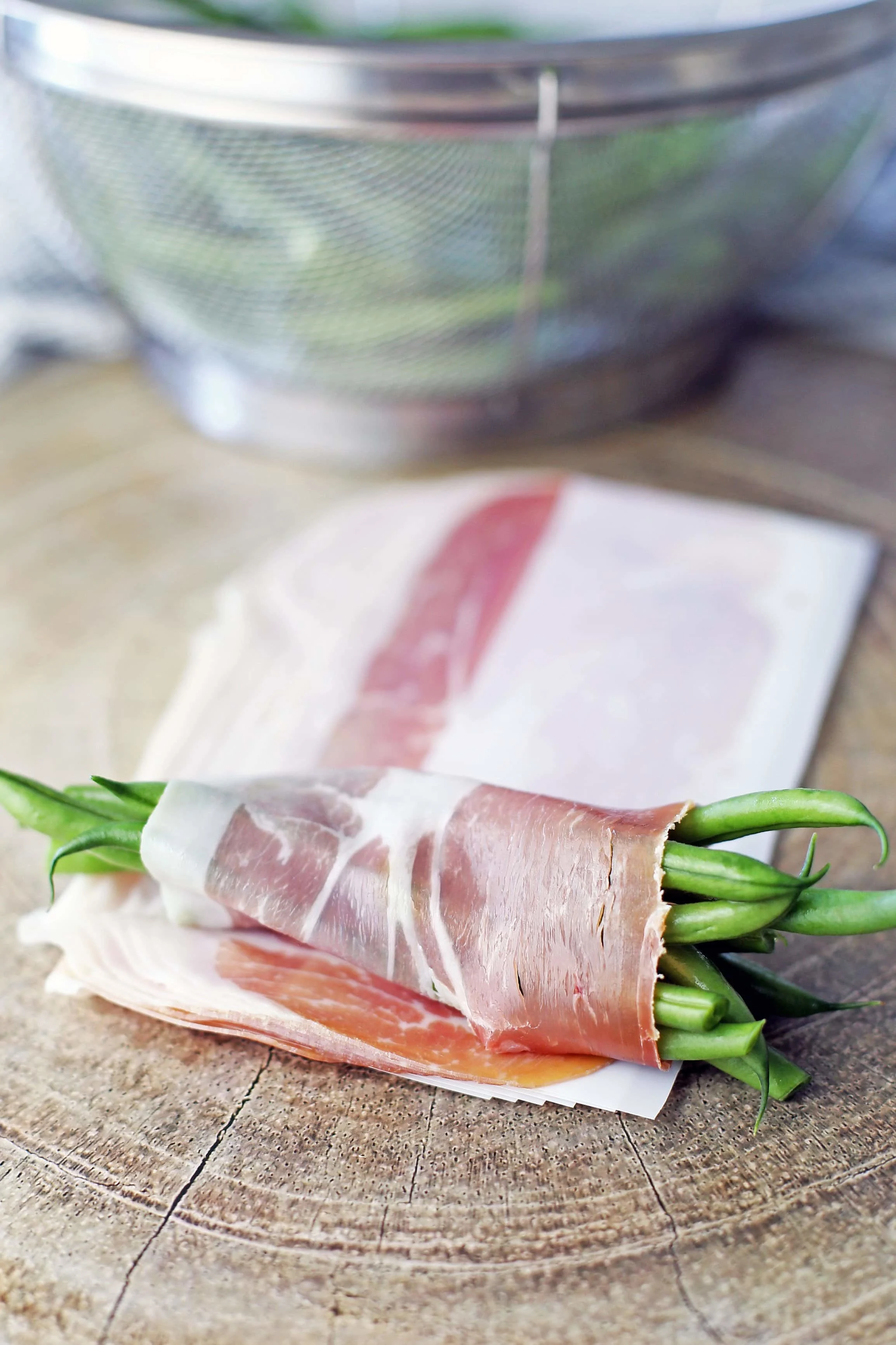 Ten French green beans wrapped in a single slice of prosciutto on a wooden board.