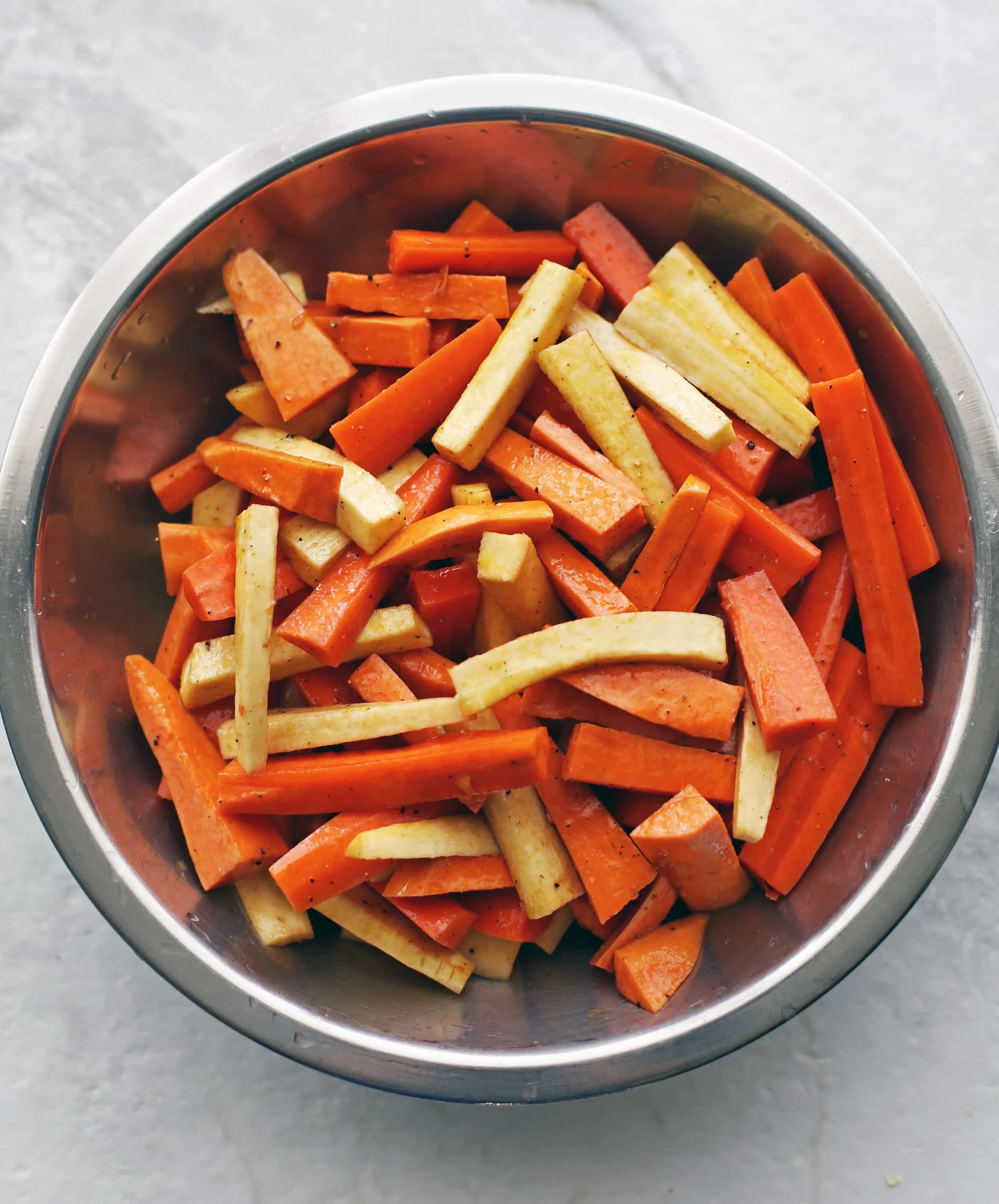 Sliced carrots, parsnips, and sweet potatoes mixed with olive oil and balsamic vinegar in a large metal bowl.