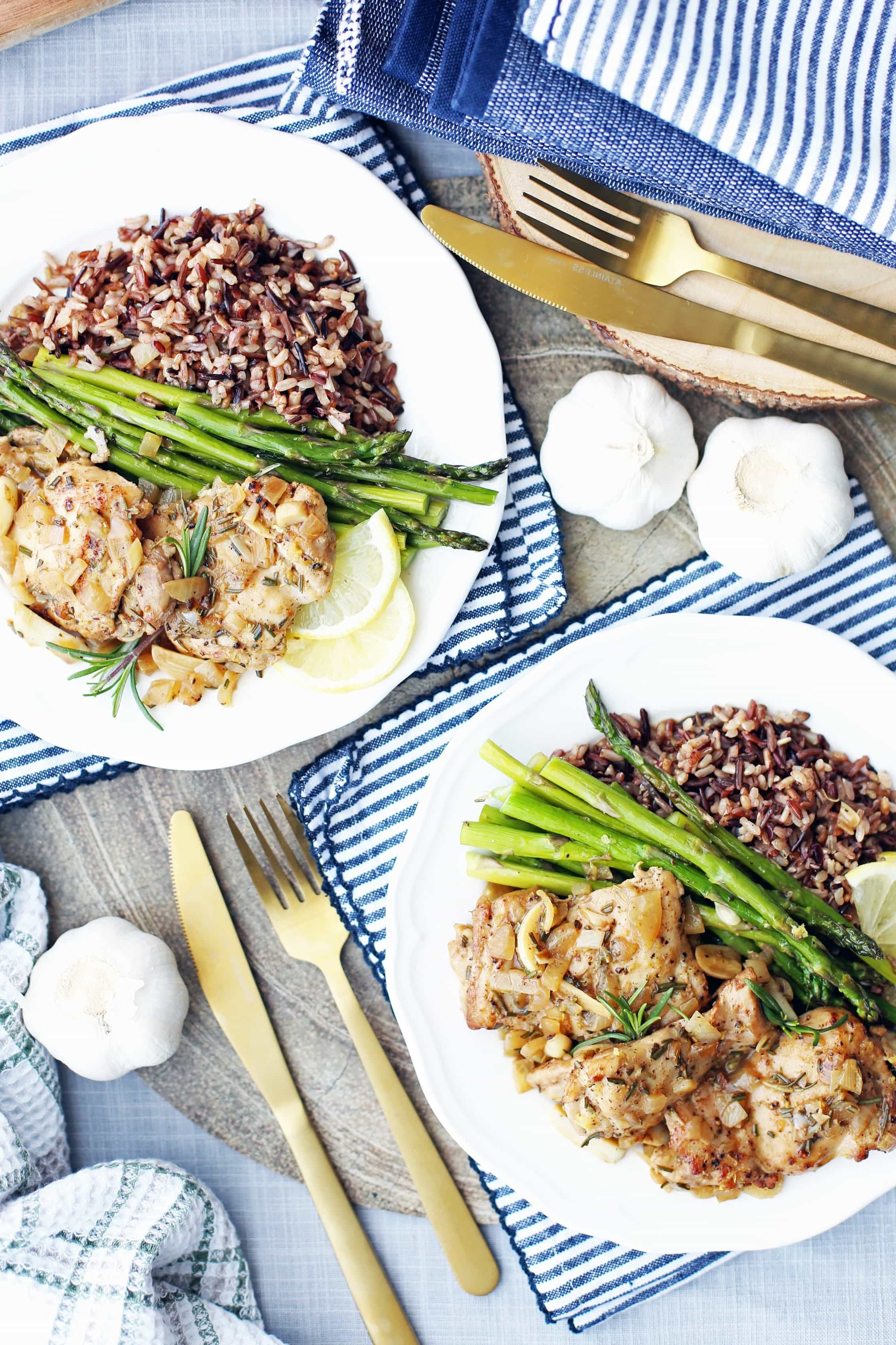 Overhead view of two white plates containing rosemary garlic chicken thighs, roasted asparagus, and wild rice.