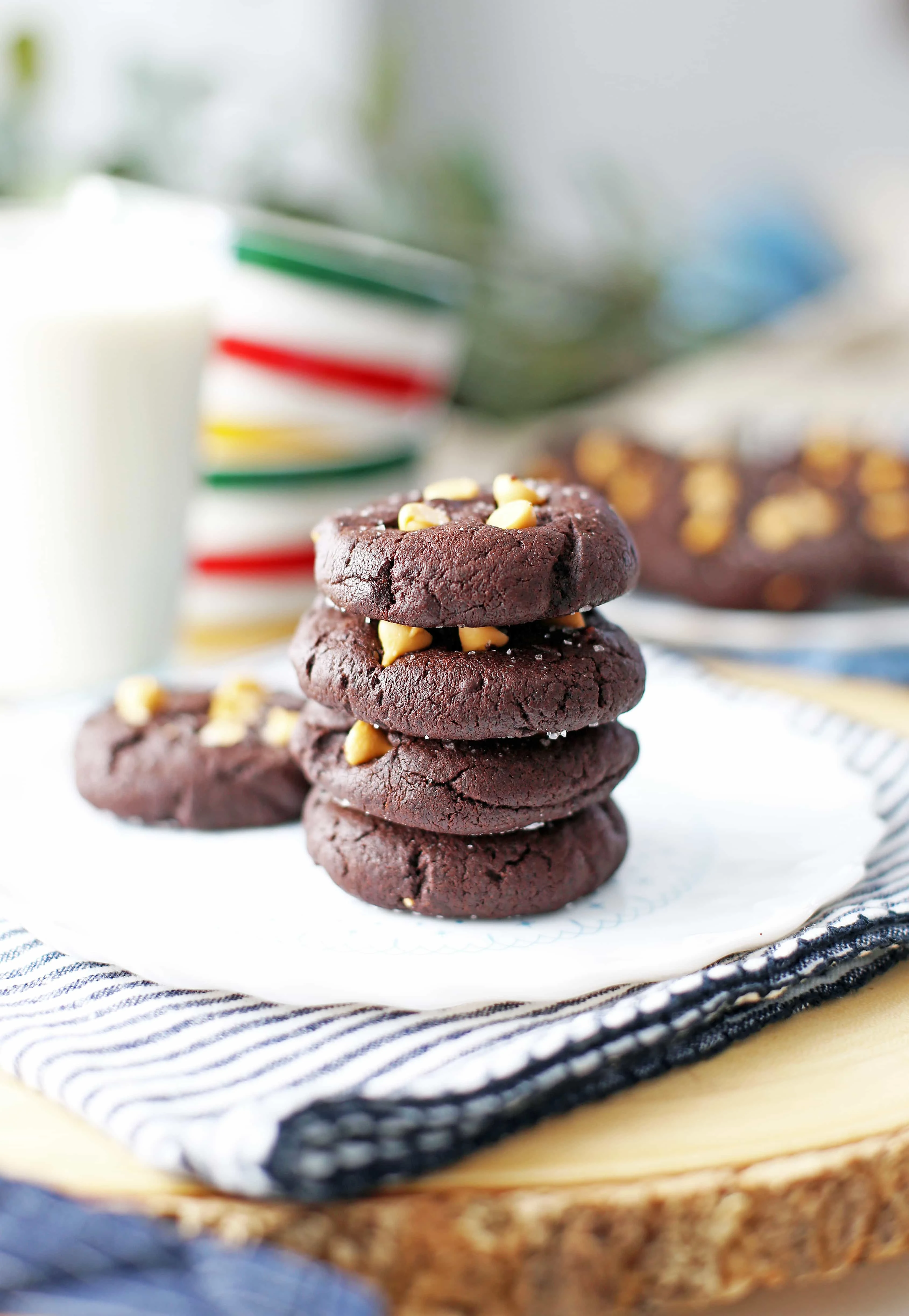 Chocolate butterscotch chip cookies stacked on a white plate with more cookies and a glass of milk behind it.