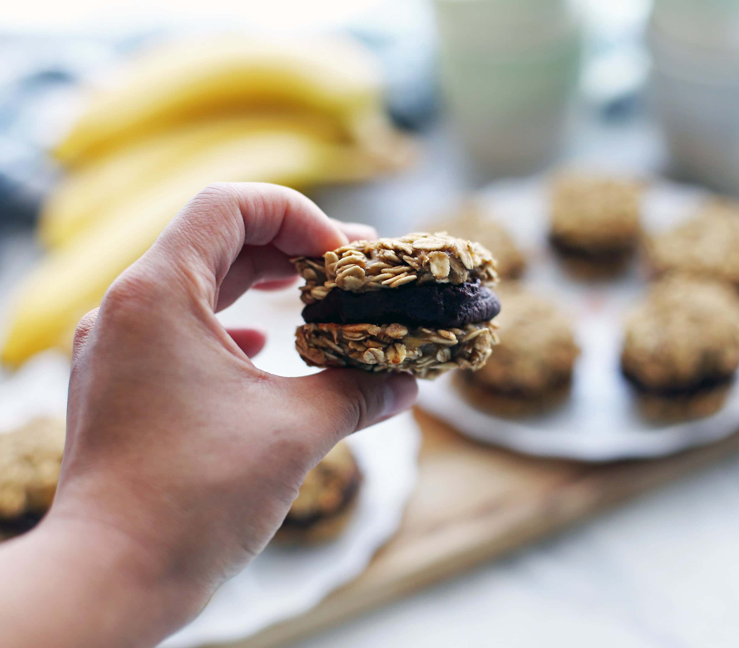 A hand holding a single banana oatmeal sandwich cookies with peanut butter cocoa filling.