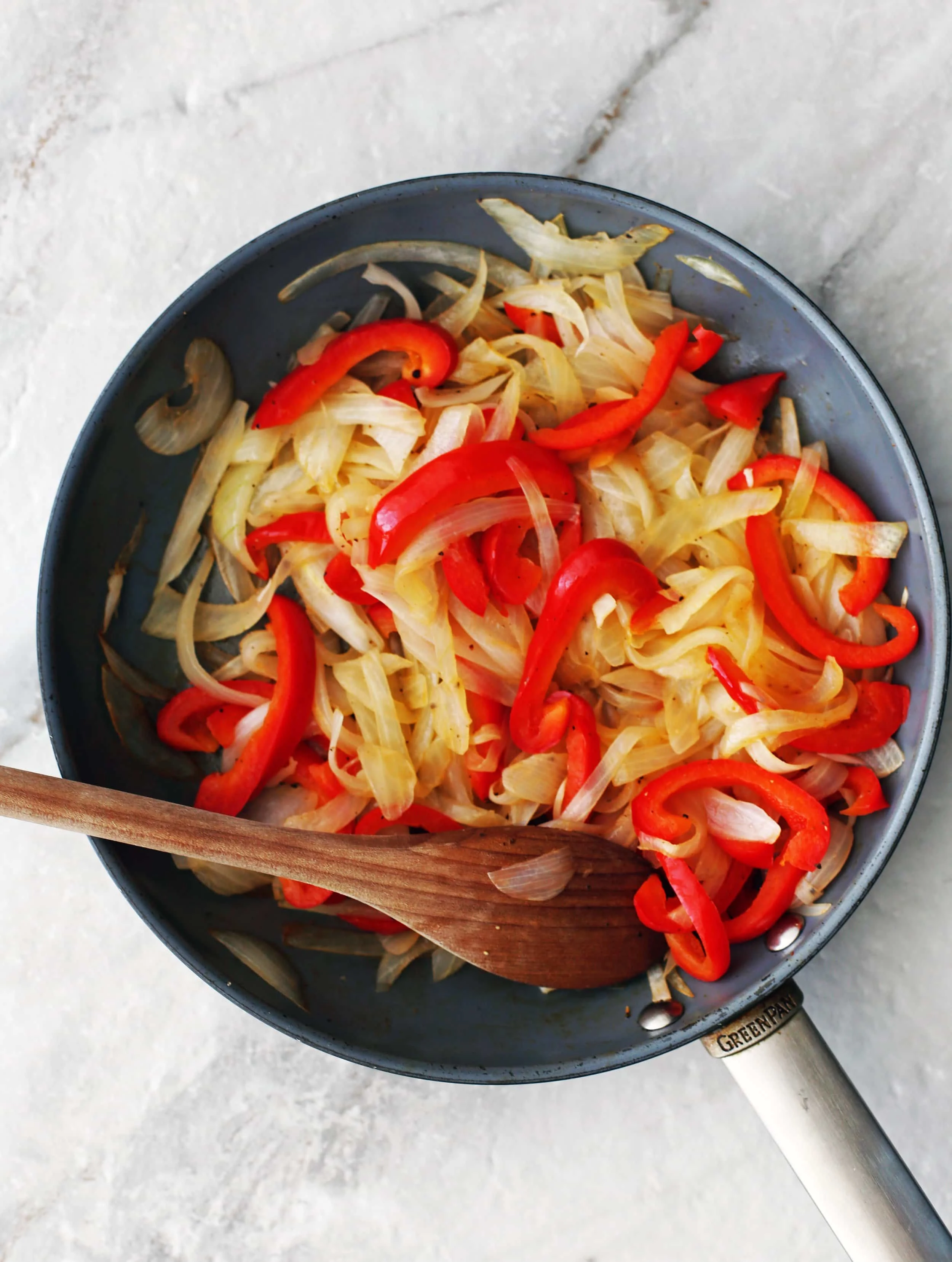 Sauteed sliced onions and peppers in a skillet with a wooden spoon.