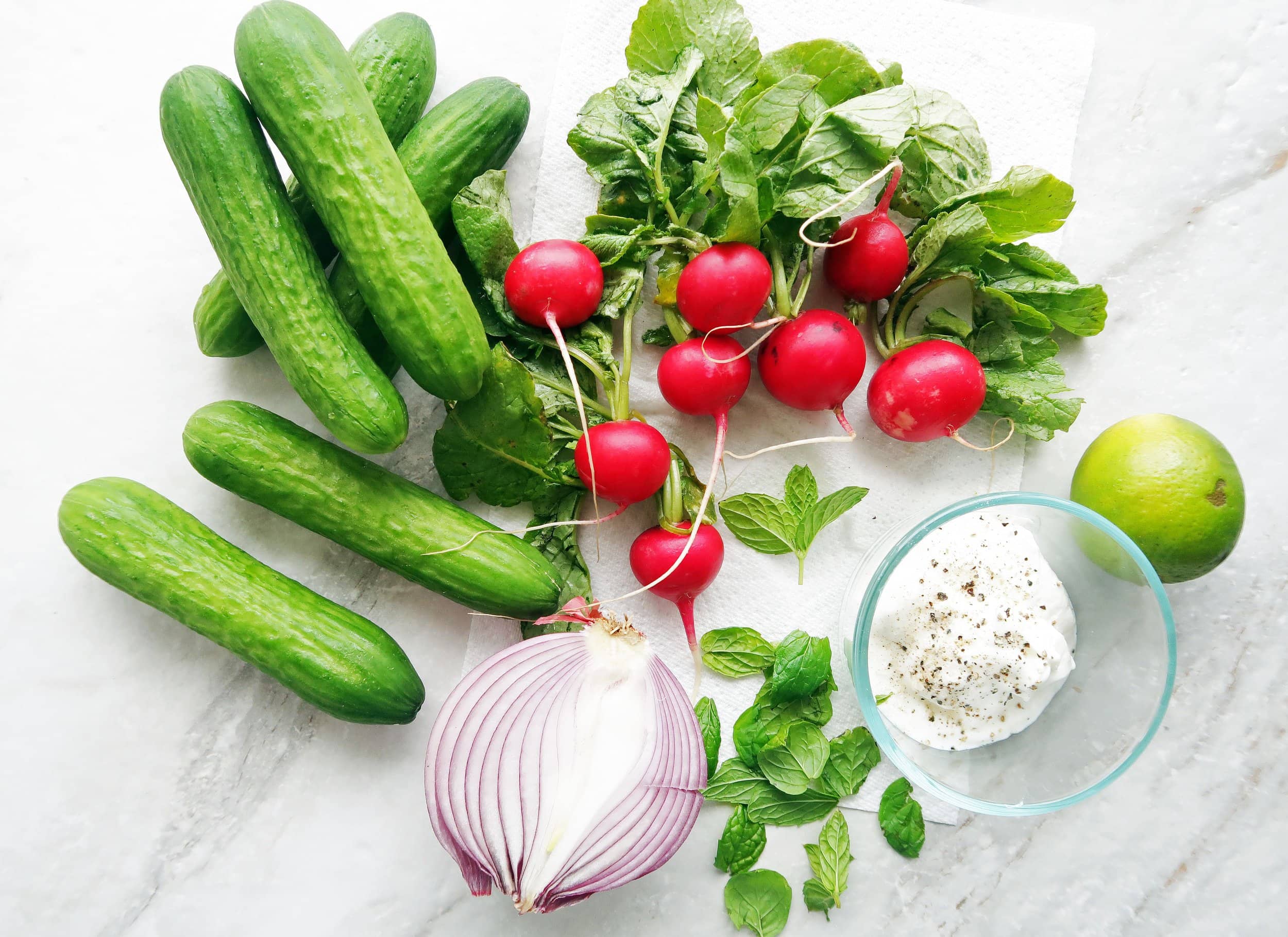 Cucumbers, radishes, a red onion, a lime, mint leaves, and a bowl of yogurt.