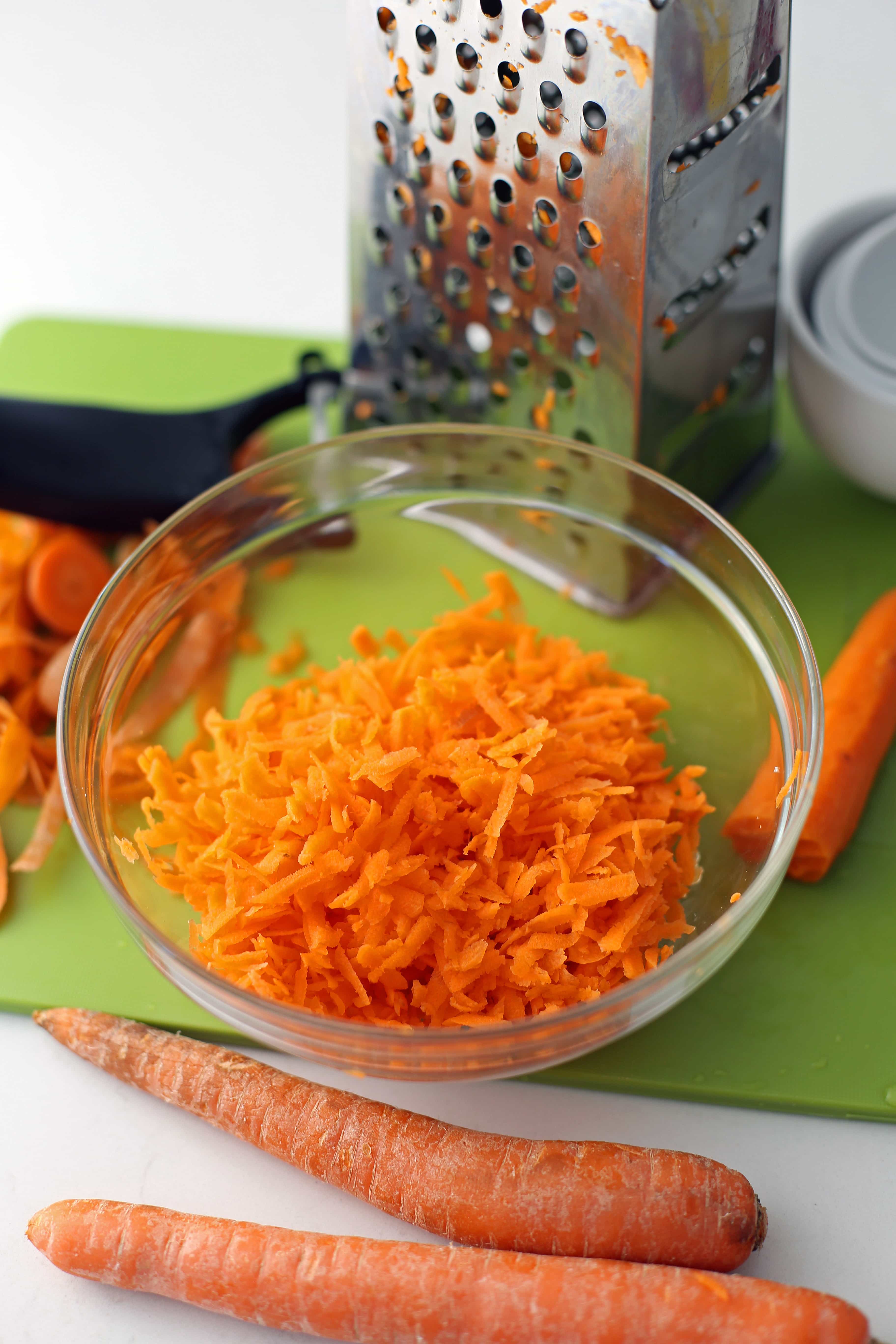 A glass bowl containing grated carrots, carrots, and metal shredder on a green cutting board.