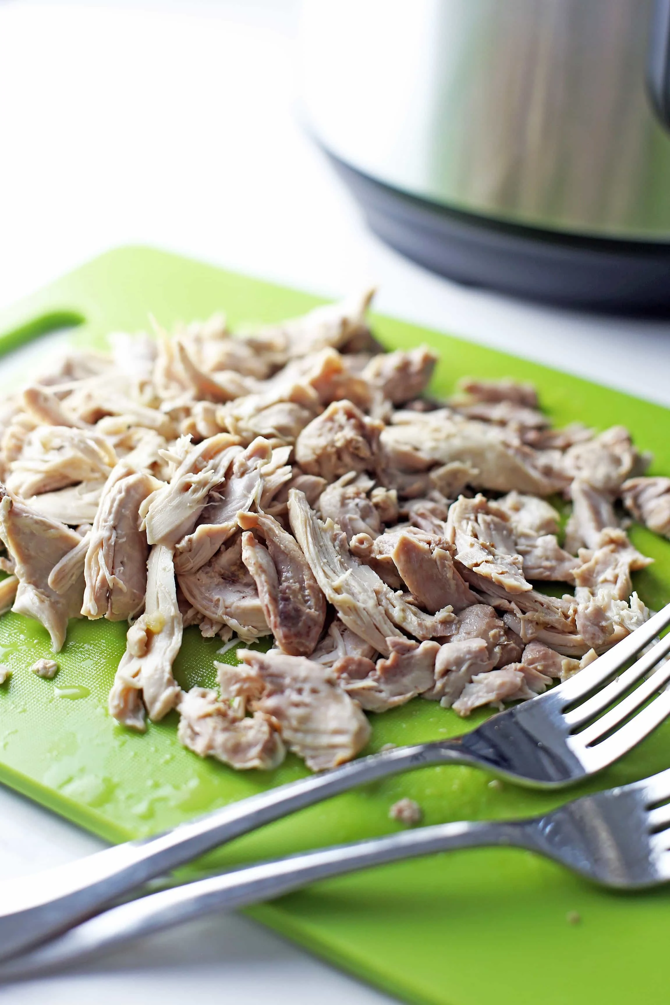 Shredded chicken thighs with two forks on top of green plastic cutting board.