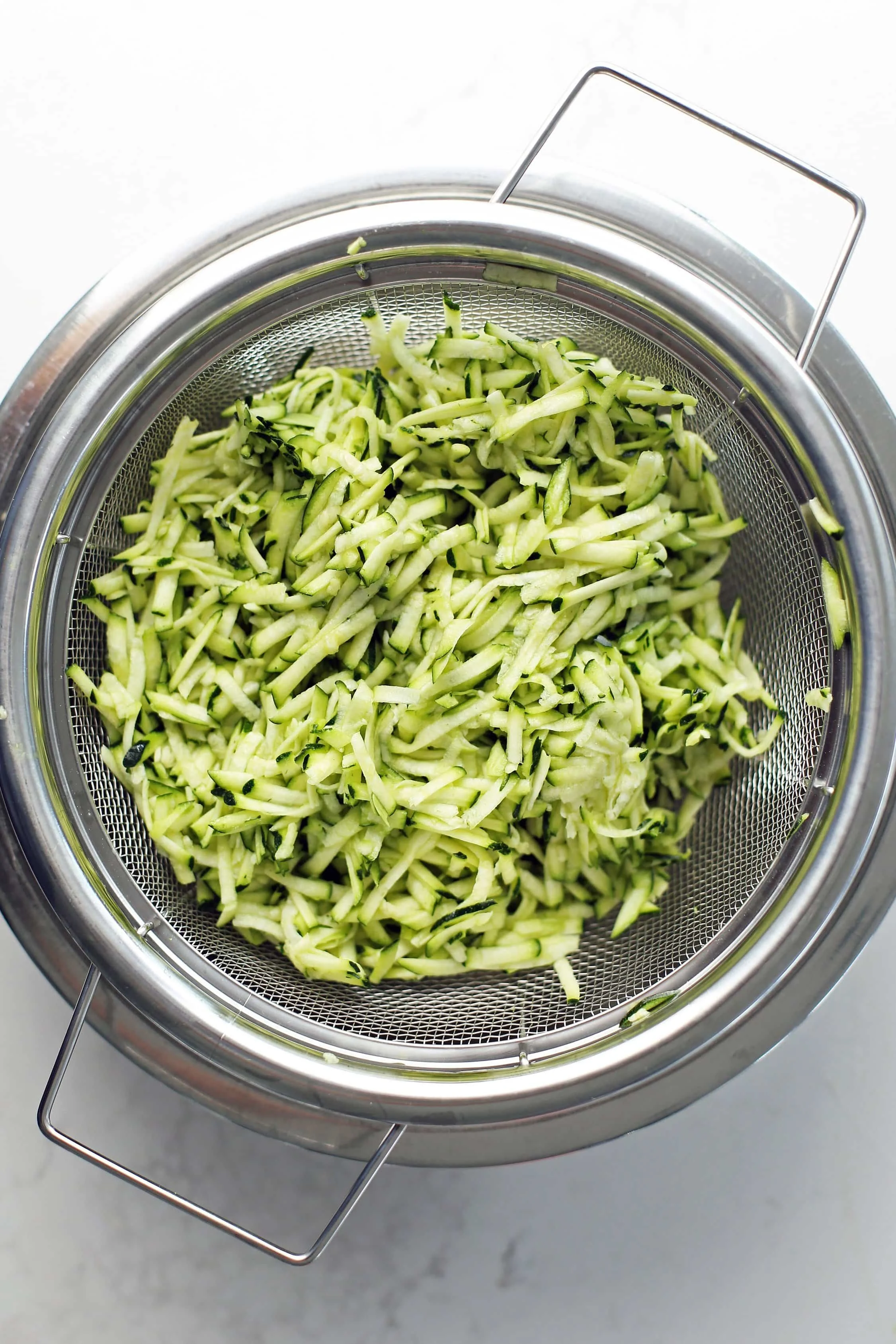 Grated zucchini in a metal mesh strainer with a stainless steel bowl placed below it.