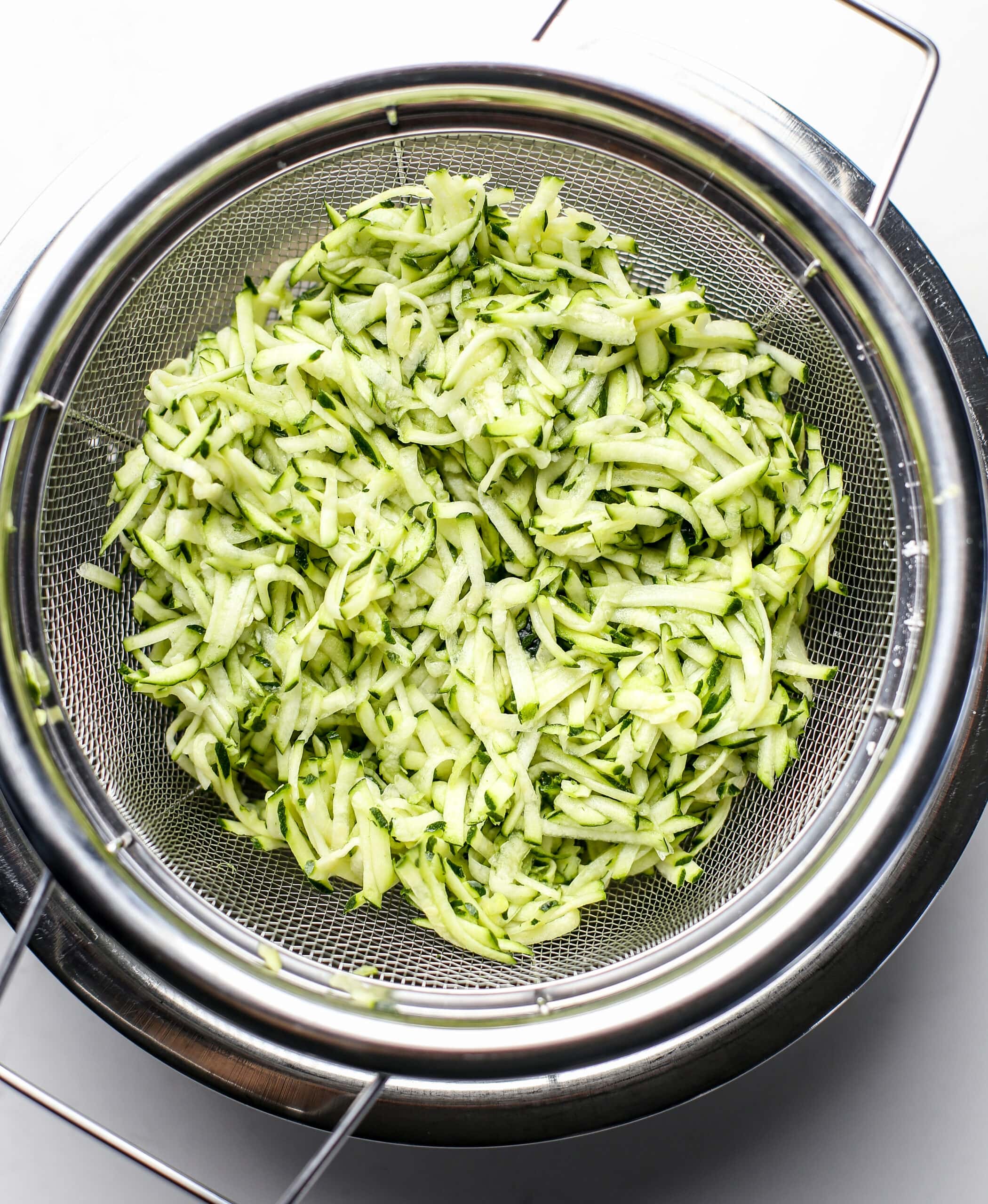Shredded zucchini in a fine mesh strainer with a stainless steel bowl underneath.