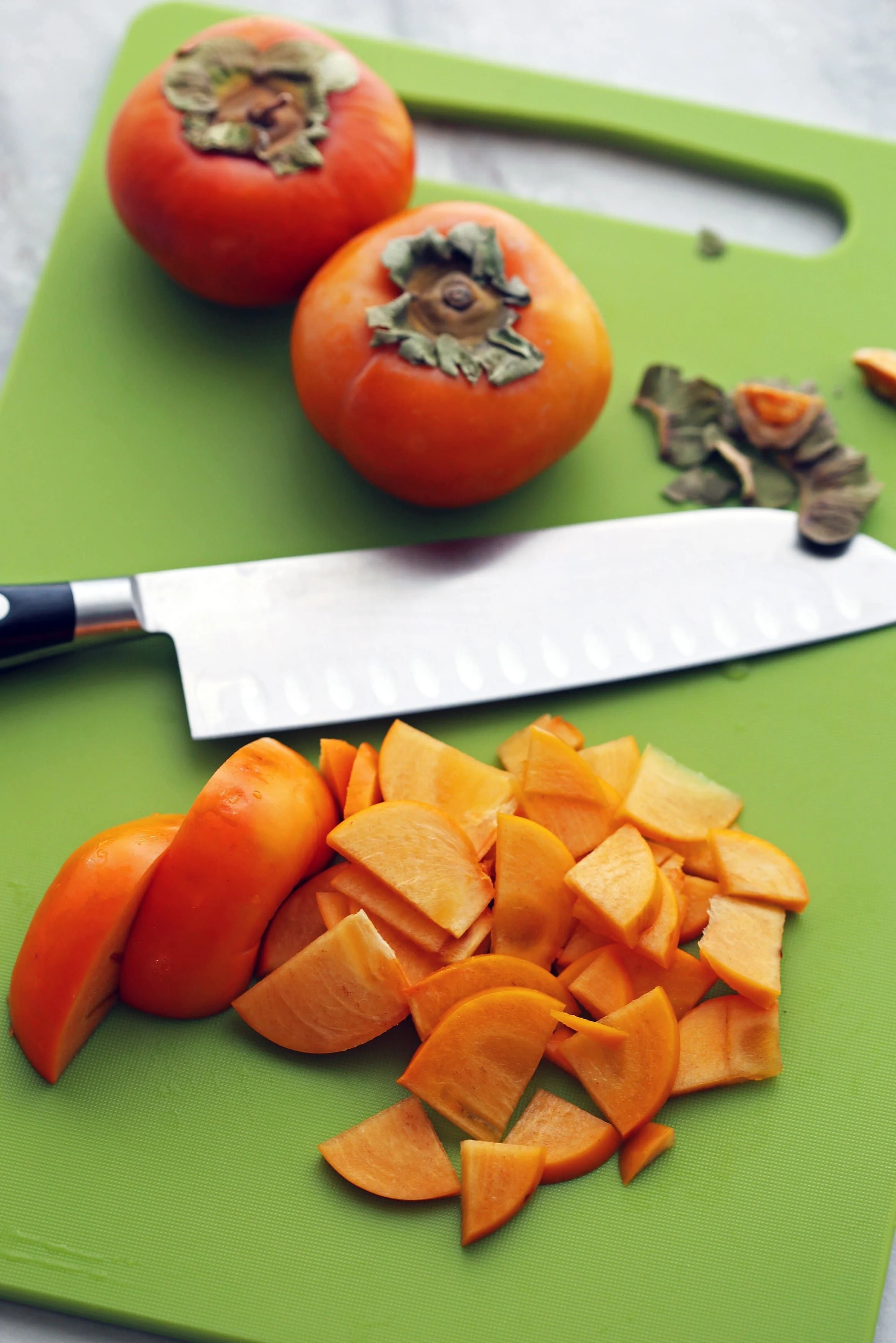 Sliced and whole persimmon fruit and a knife on a green cutting board.