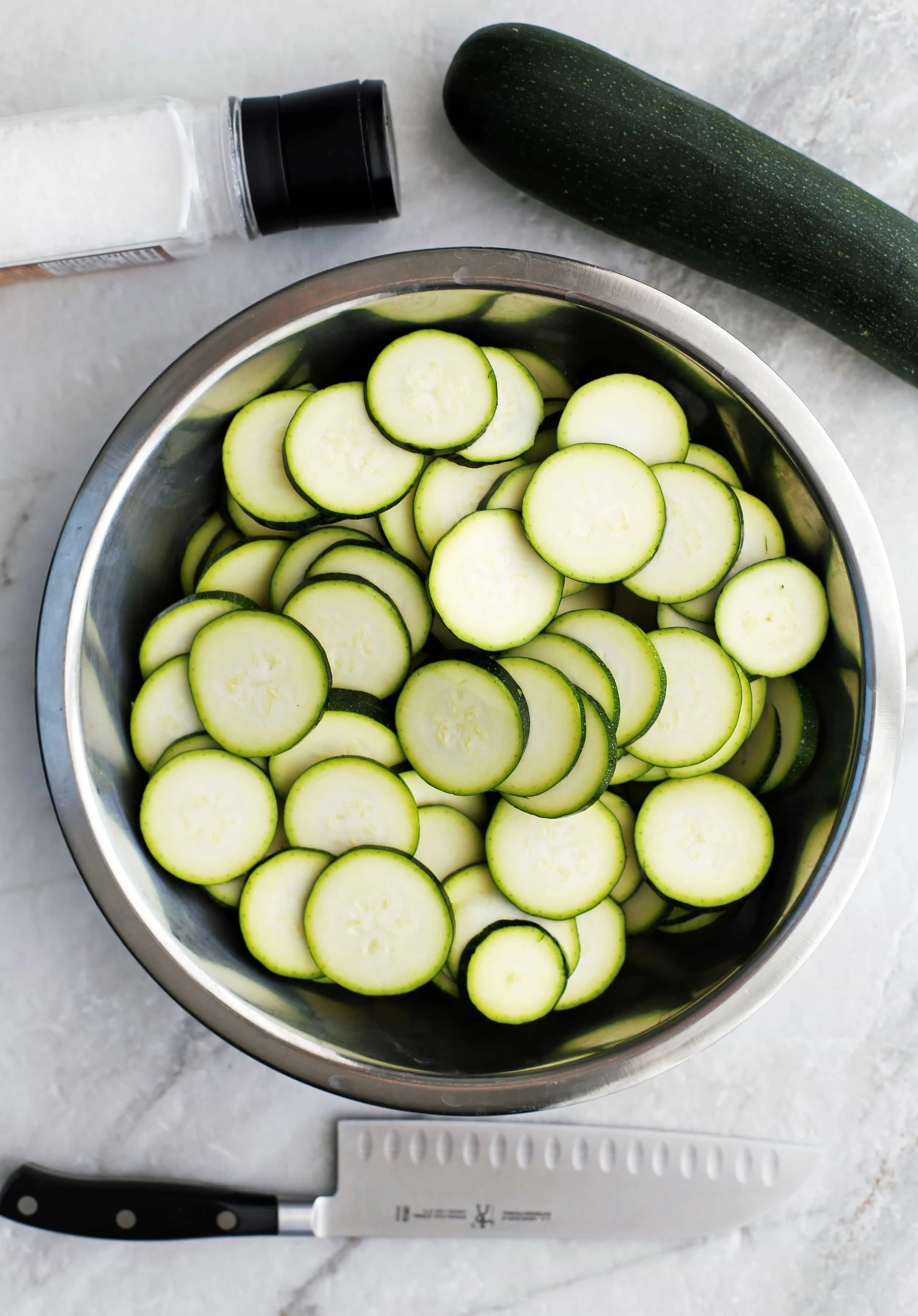 Sliced zucchini in a large metal bowl with a knife, zucchini, and salt shaker around it.