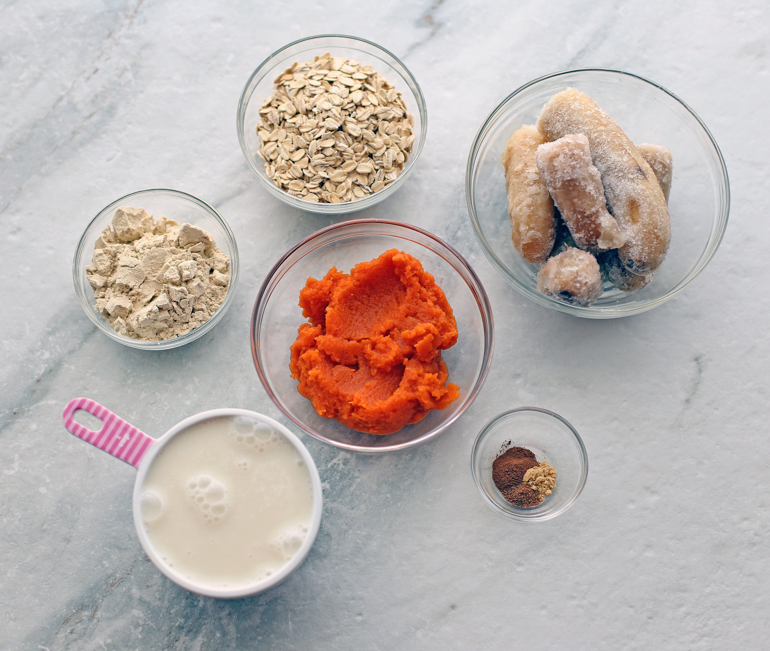 Overhead view of almond milk, pumpkin puree, frozen banana, spices, protein powder, and oatmeal.