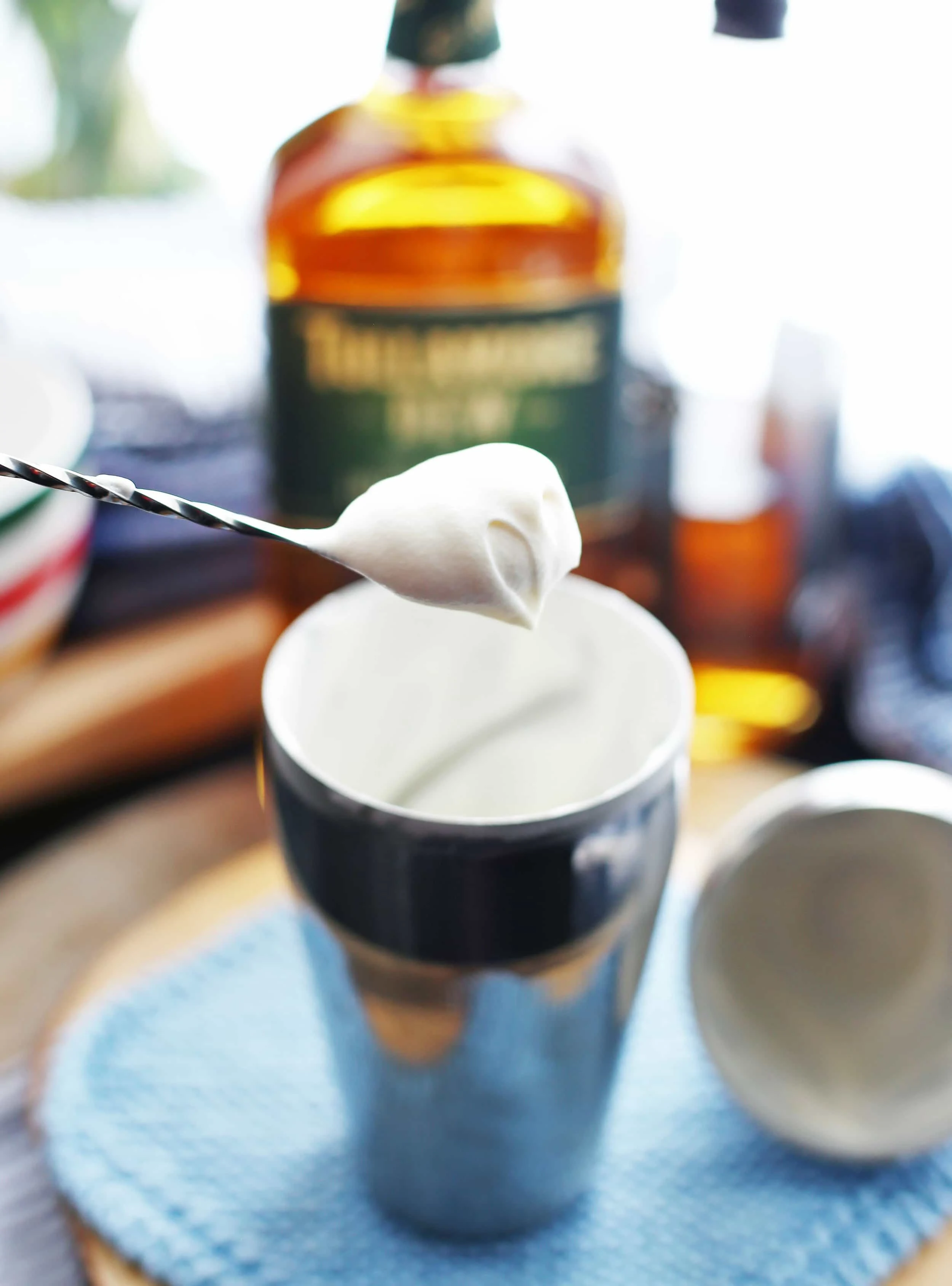 A long cocktail spoon holding a dollop of lightly whipped cream over a stainless steel cocktail shaker.