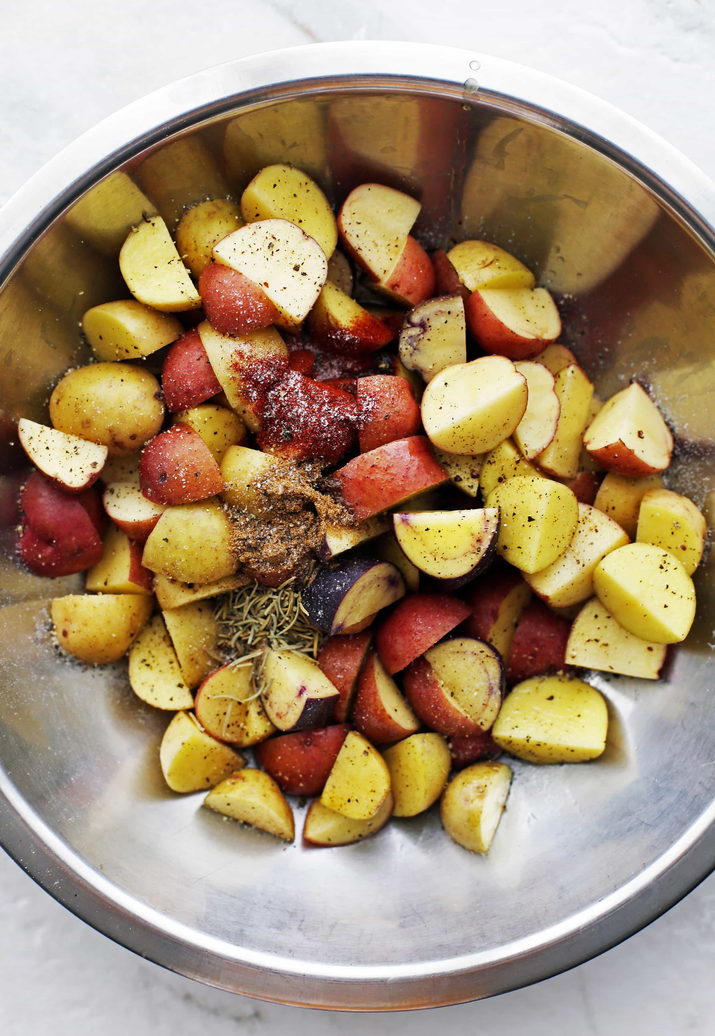 Quartered baby potatoes, olive oil, spices, and dried rosemary in a metal bowl.