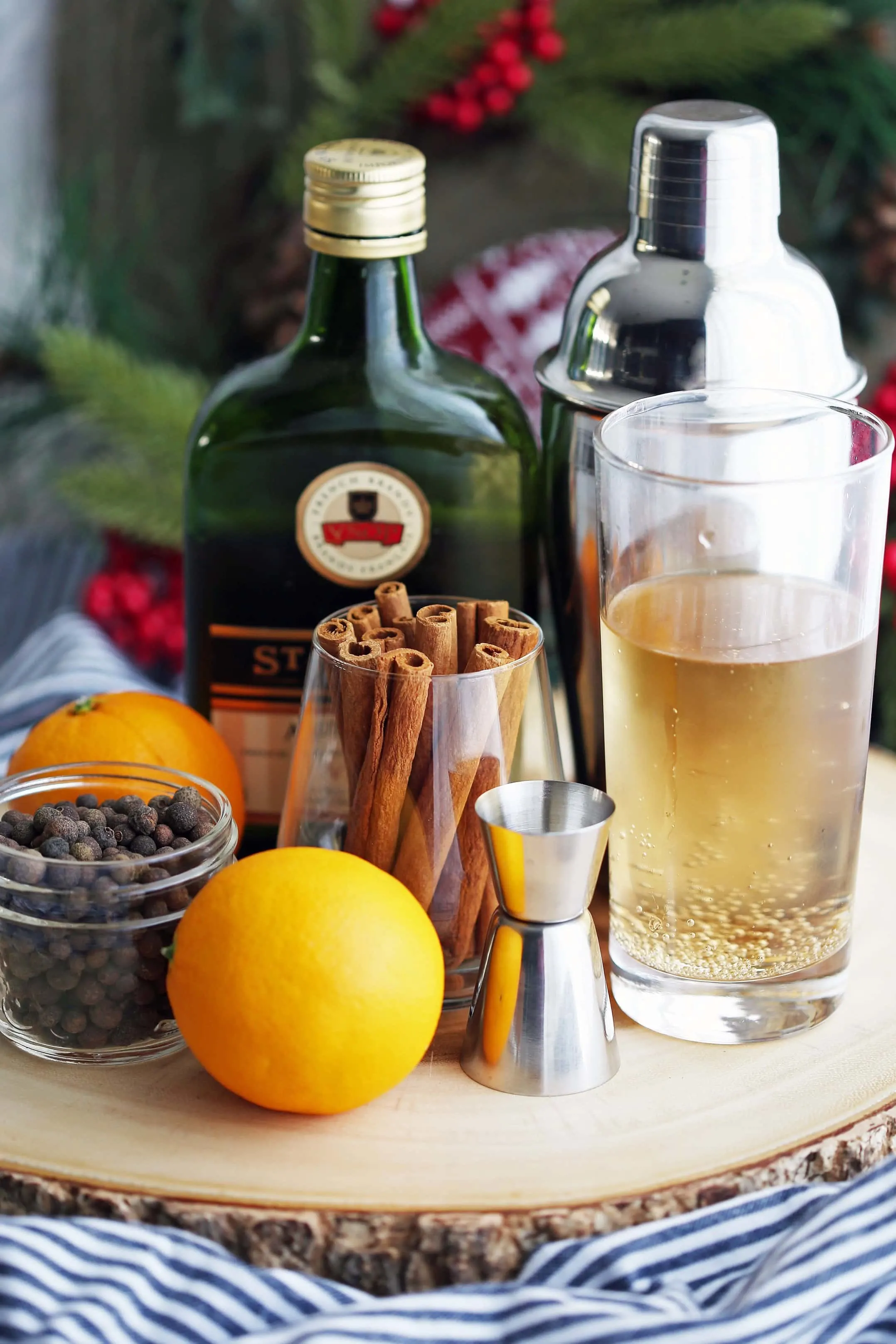 Whole allspice berries, cinnamon sticks, a bottle of Brandy, an orange, a glass of ginger ale, and a cocktail shaker on a wooden platter.