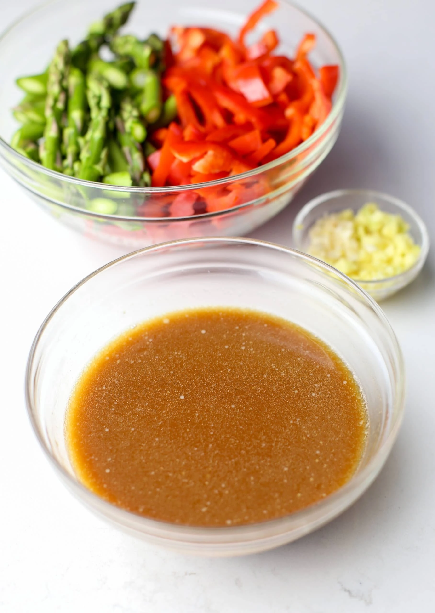 Stir-fry sauce, minced ginger and garlic, and sliced asparagus and bell pepper in three separate glass bowls.