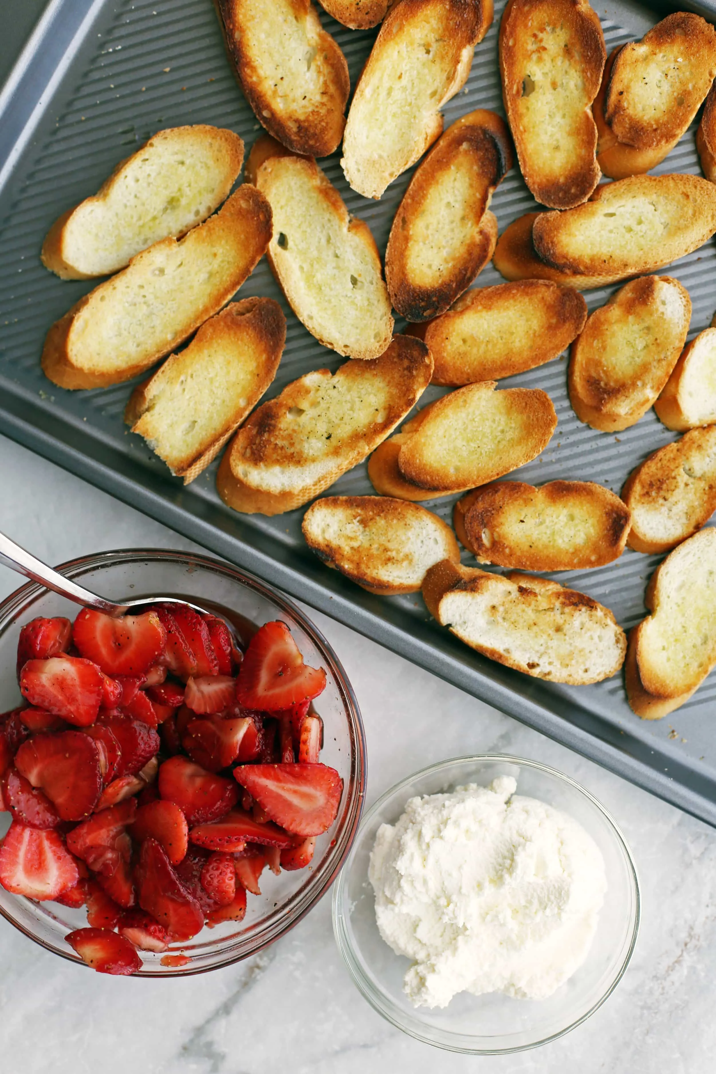 Toasted baguette slices on a baking sheet, a bowl of balsamic strawberries, and a bowl of ricotta.