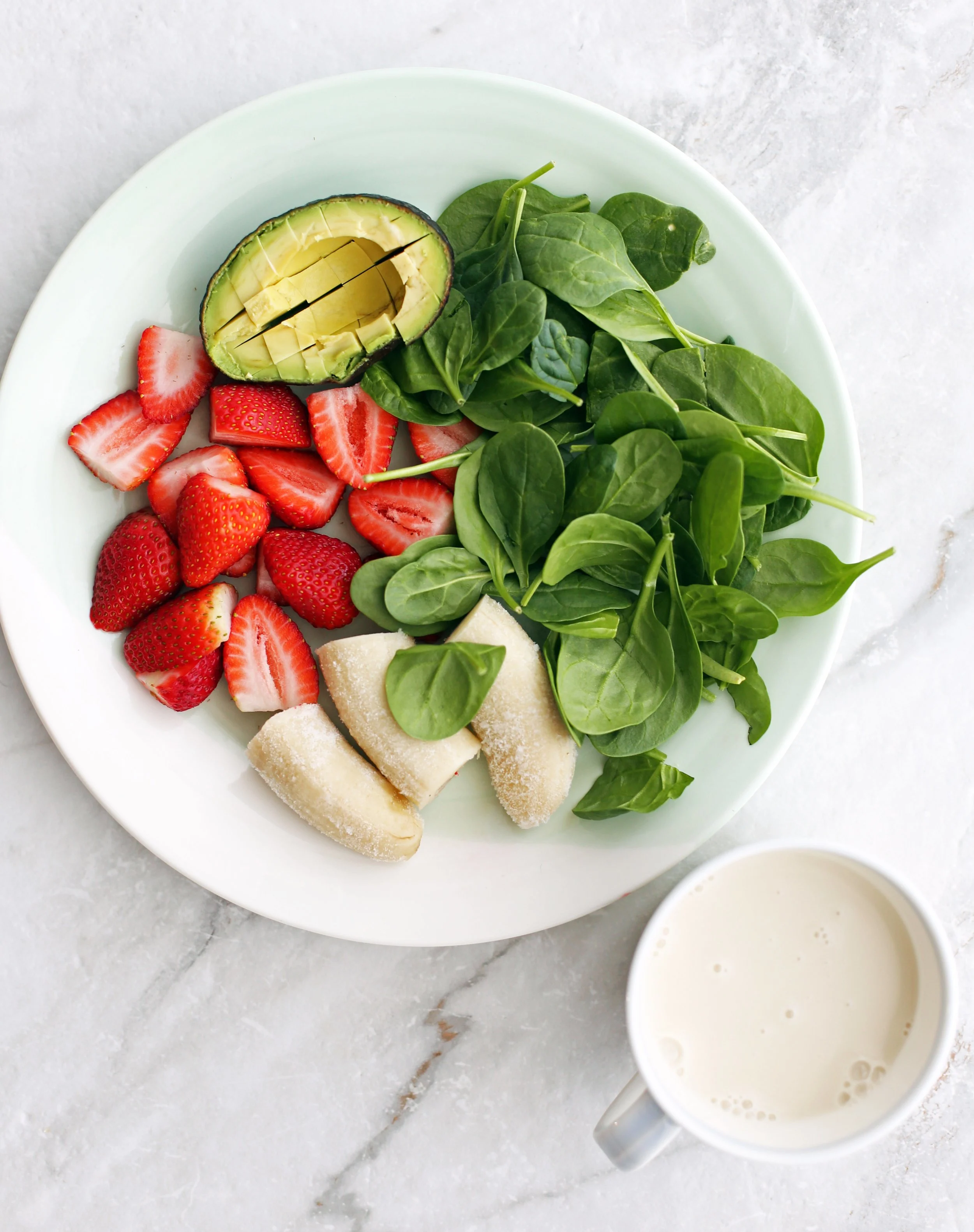 A plate of sliced strawberries, frozen banana, baby spinach, and half avocado with a cup of almond milk.