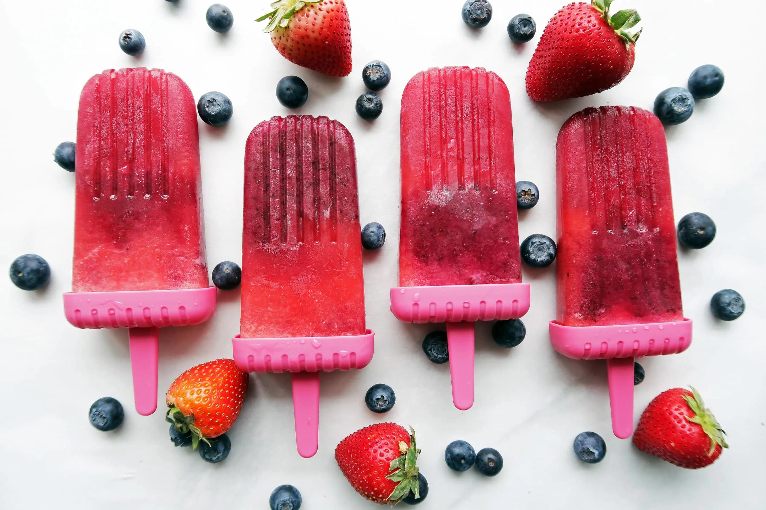 Four Strawberry Blueberry Coconut Water Popsicles placed side by side with strawberries and blueberries around them. 