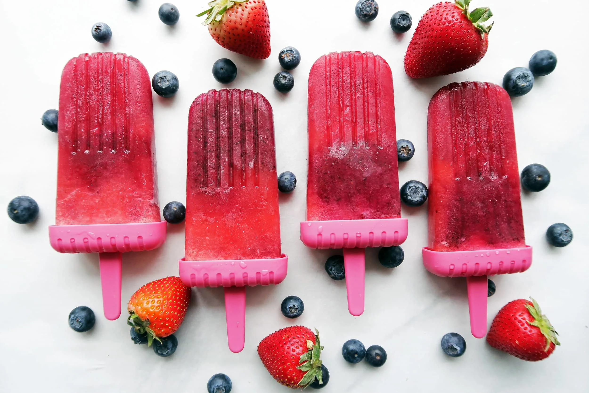 Four Strawberry Blueberry Coconut Water Popsicles placed side by side with strawberries and blueberries around them.