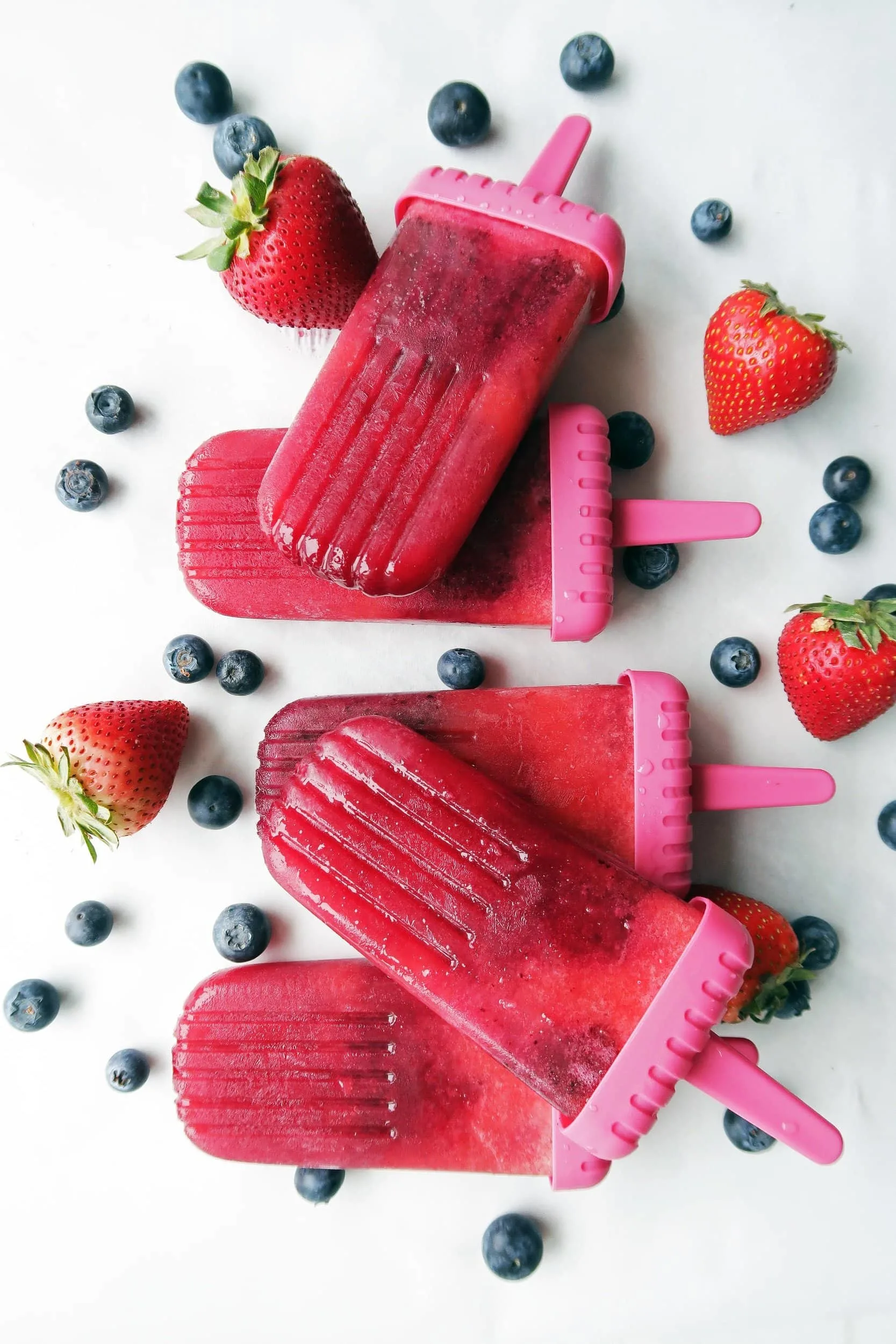 Five Strawberry Blueberry Coconut Water Popsicles with strawberries and blueberries around them.Strawberry Blueberry Coconut Water Popsicles placed beside and overlapping one another with strawberries and blueberries around them.