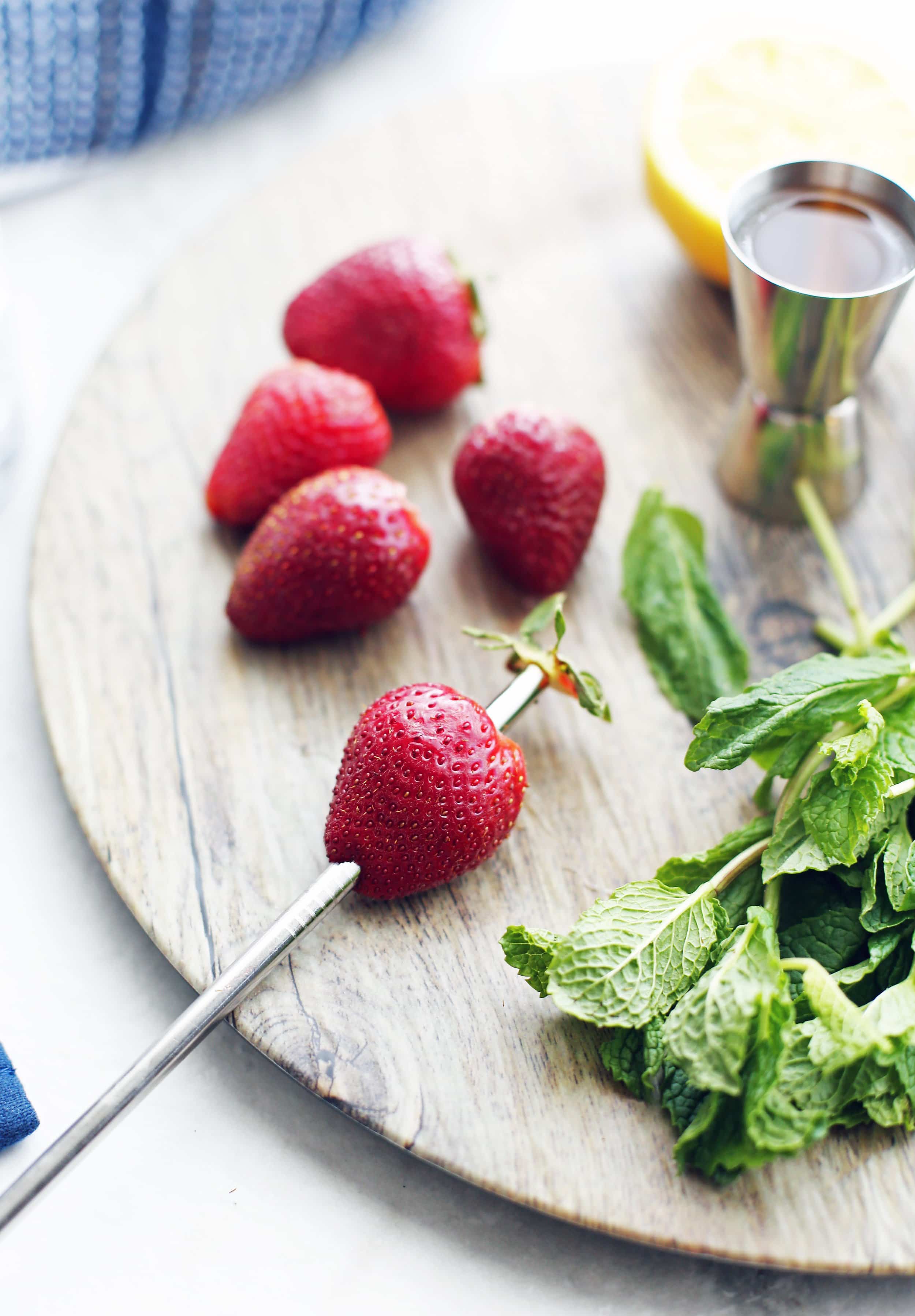 A metal straw pierced through a strawberry with the stem pushed out from the straw. Fresh mint and hulled strawberries beside it.