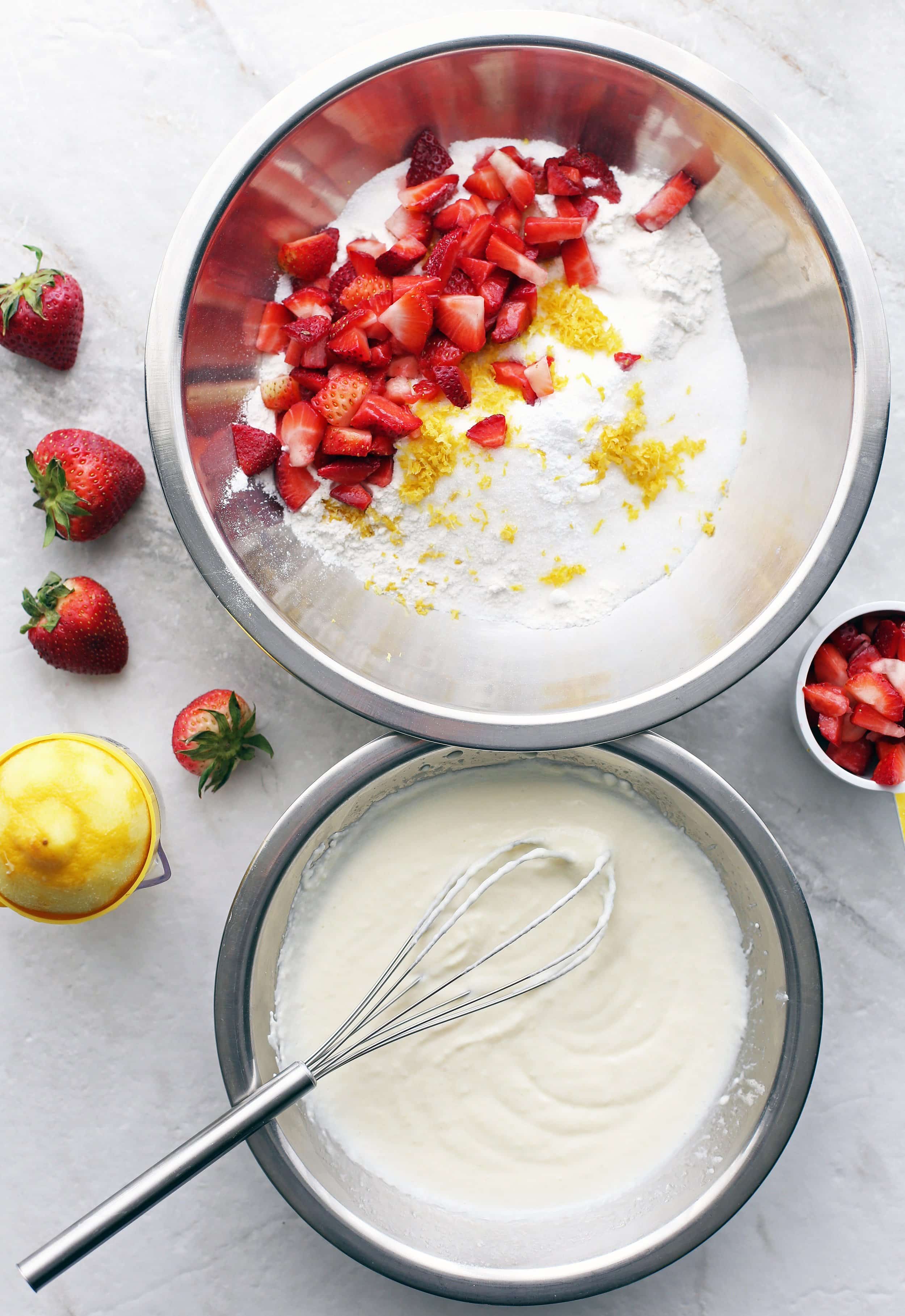 One metal bowl containing dry baking ingredients, lemon zest, and strawberries. Another bowl containing whisked wet ingredients.