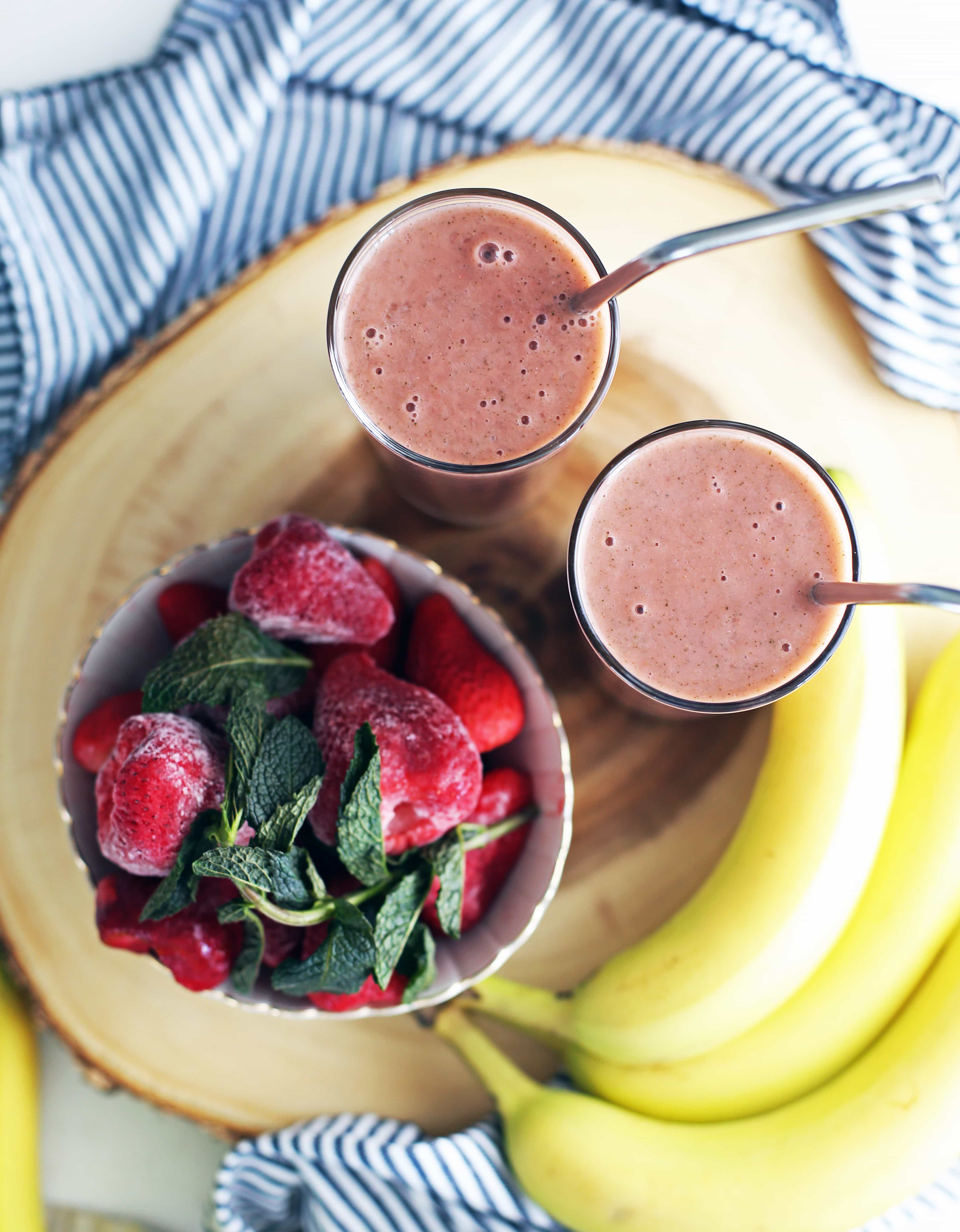 Overhead view of two glasses full of strawberry mint smoothie, a bowl of strawberries and mint, and bananas on the side. 