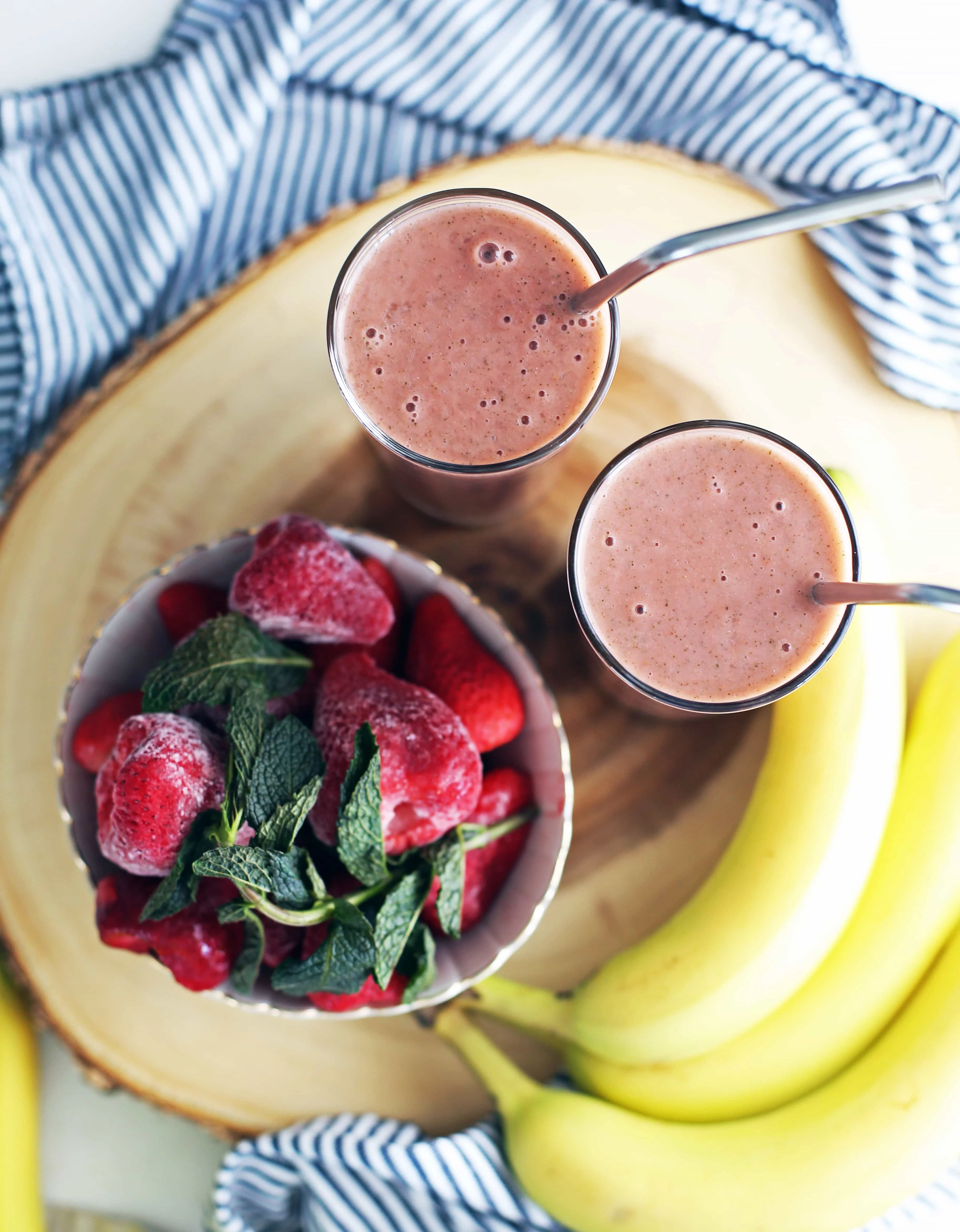 Overhead view of two glasses full of strawberry mint smoothie, a bowl of strawberries and mint, and bananas on the side. 