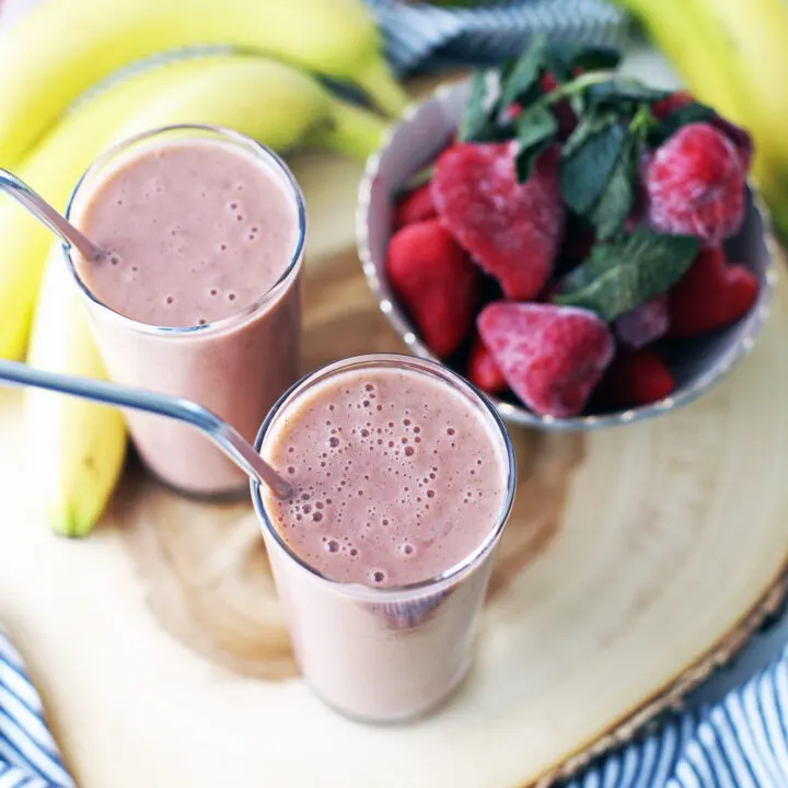 Two strawberry mint smoothies, a bowl of frozen strawberries, and bananas on a wooden platter.