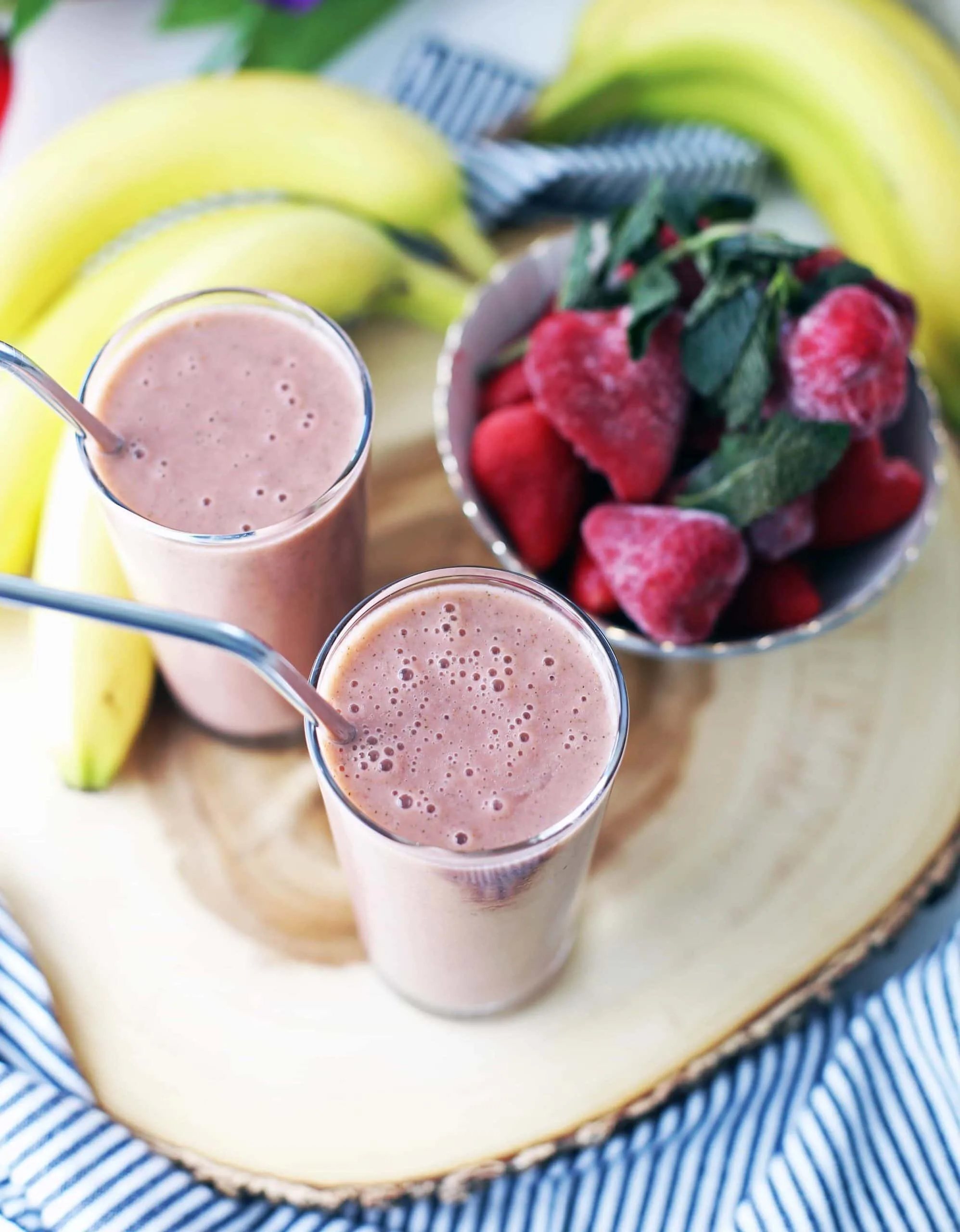 Two strawberry mint smoothies, a bowl of frozen strawberries, and bananas on a wooden platter.