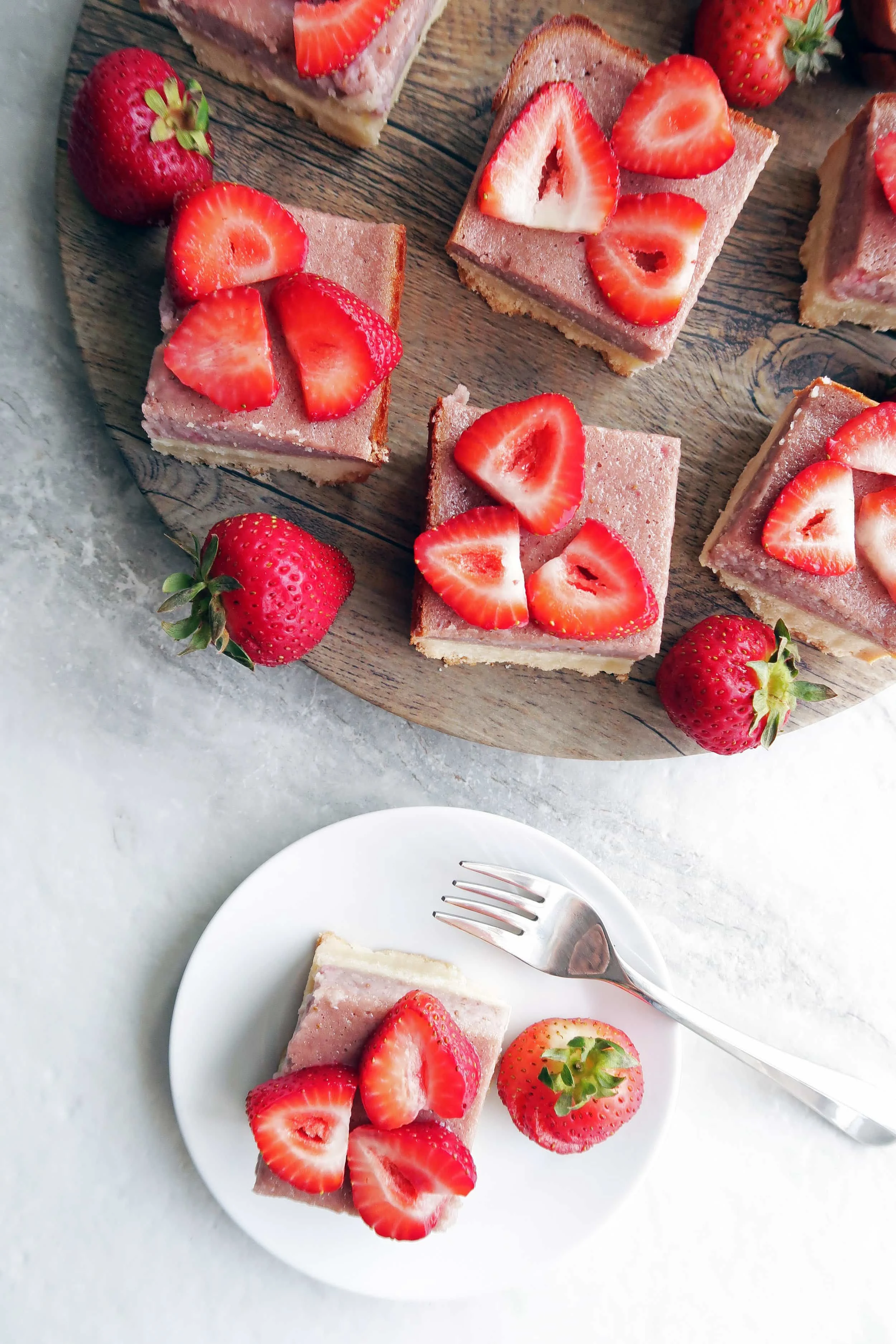 Pink strawberry shortbread bars with fresh strawberry slices on top on a round wooden platter and on a small white plate.