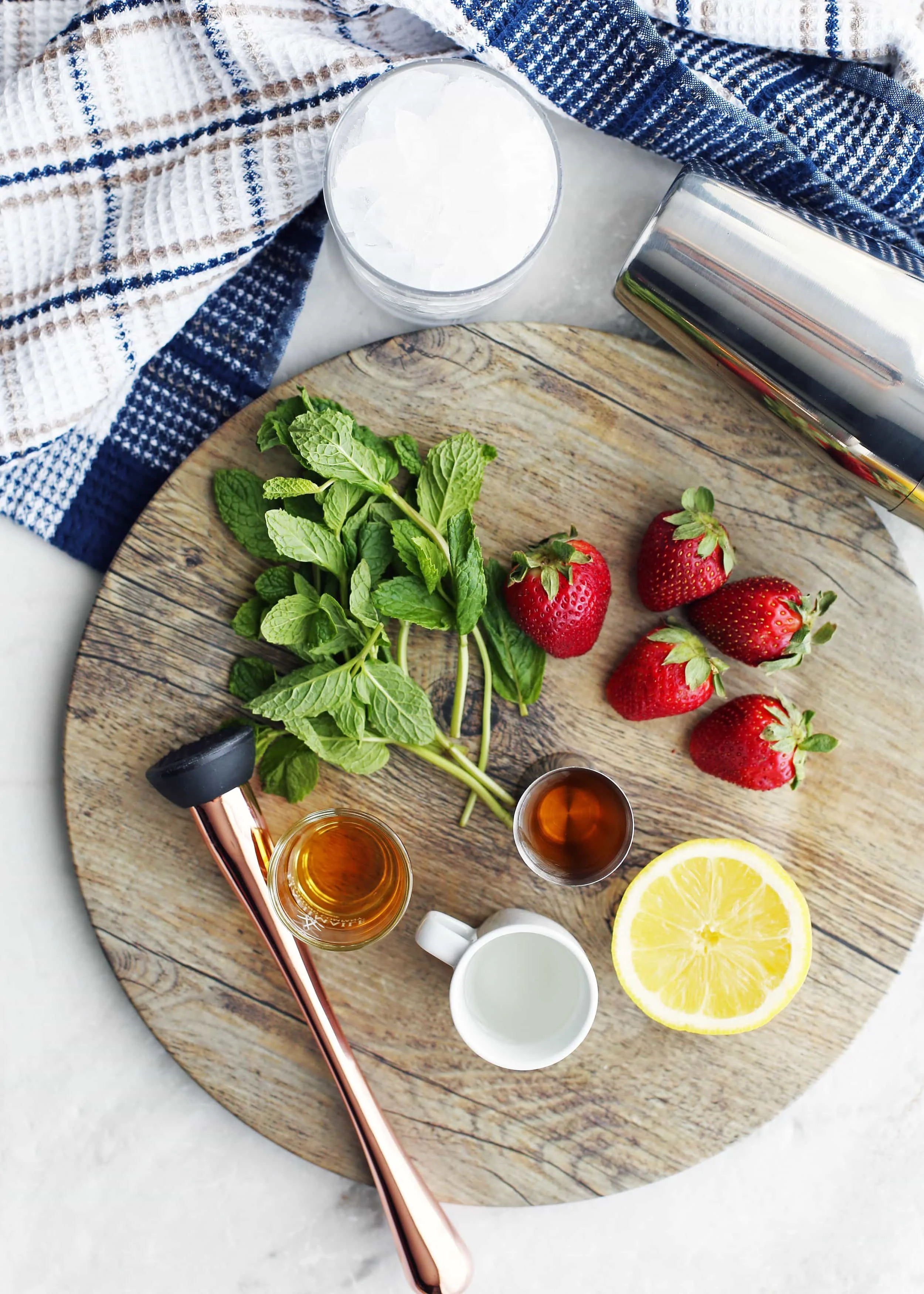 A wooden plate containing fresh mint, strawberries, lemon, small cups of whisky and simple syrup, and a muddler.