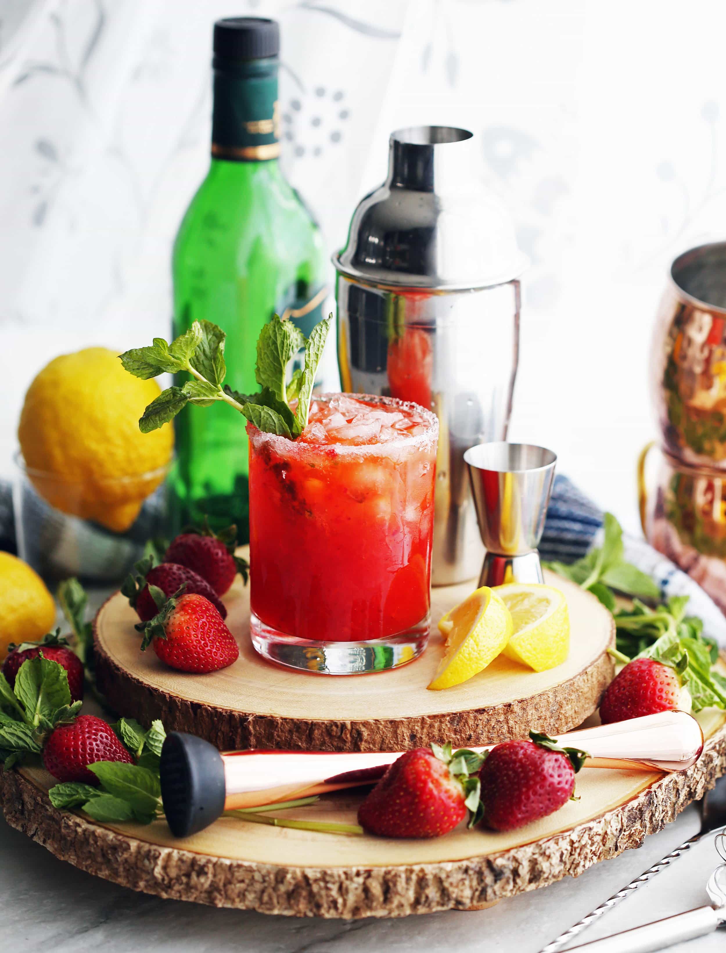A glass of Mint Strawberry Whisky Smash drink that's sitting on two wooden platters and surrounded by a cocktail set, lemons, and strawberries.