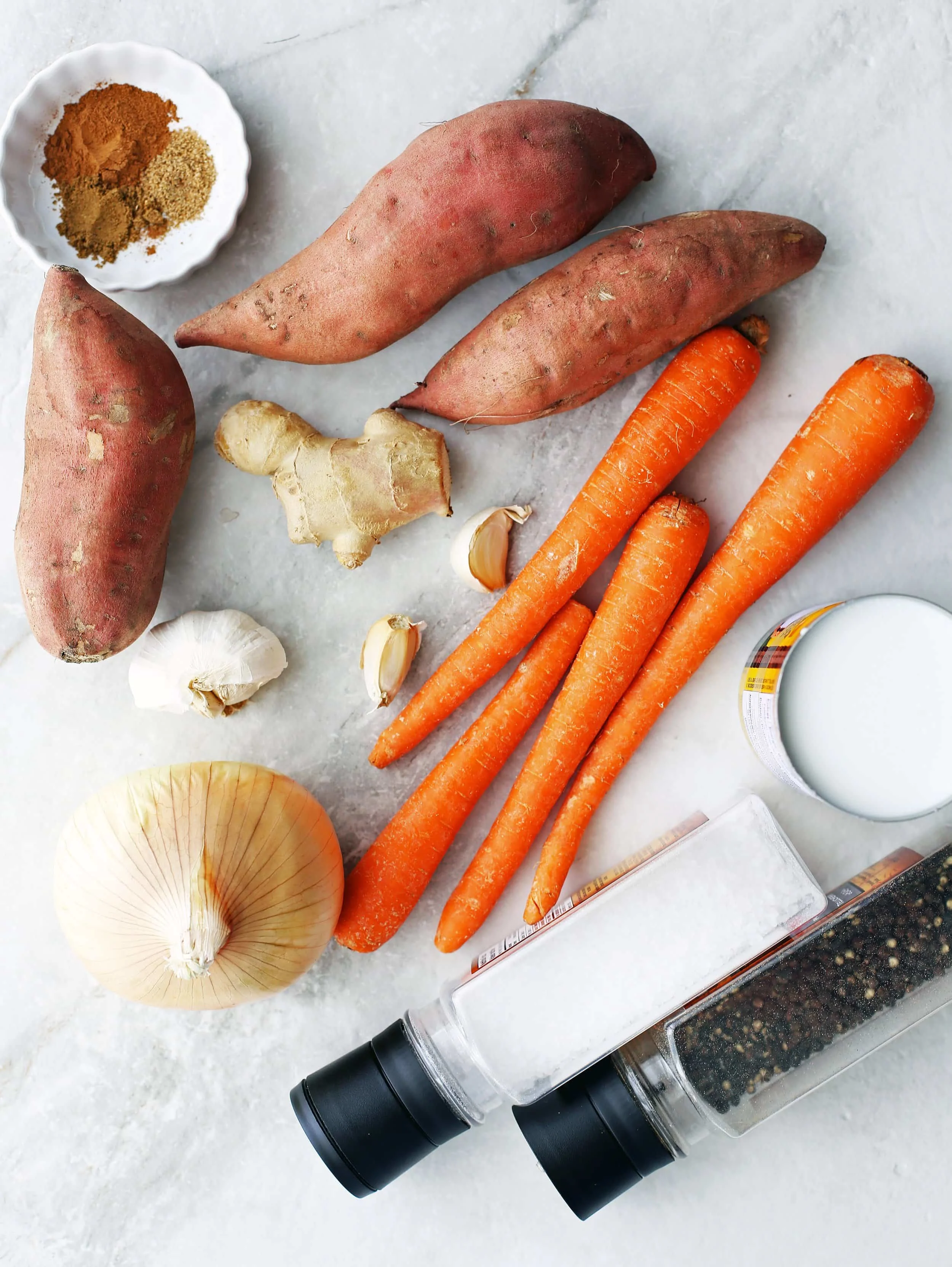 Sweet potatoes, carrots, garlic, onion, ginger, coconut milk, and spices on a white and grey surface.