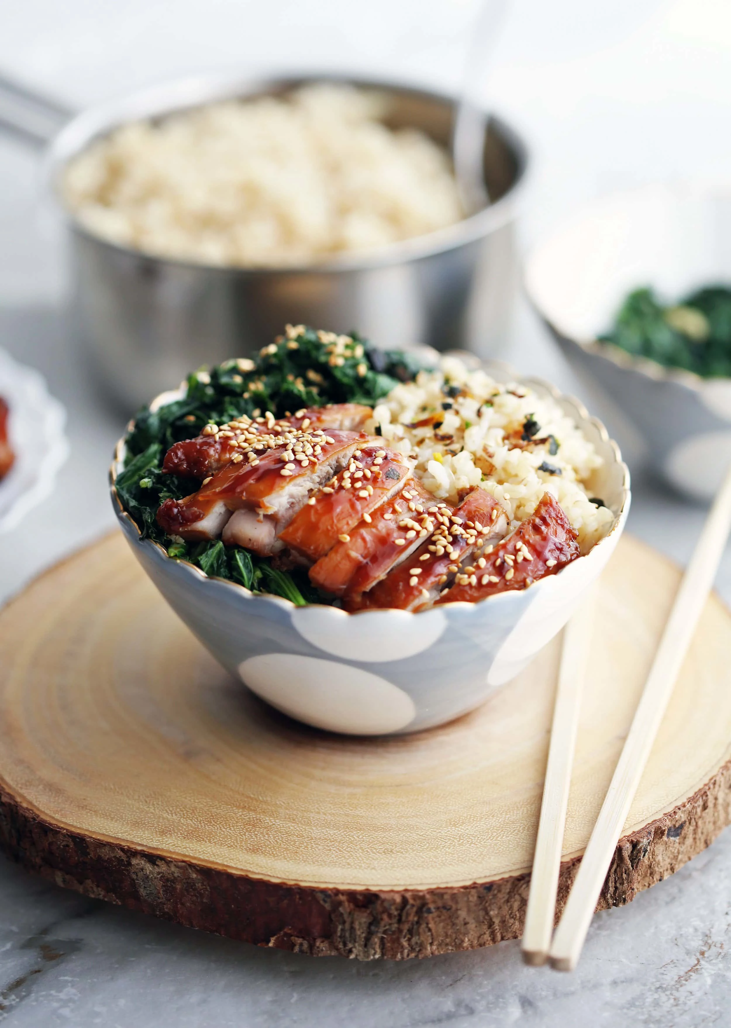 Side view of a teriyaki chicken rice bowl with garlicky kale with chopsticks on a wood slab.