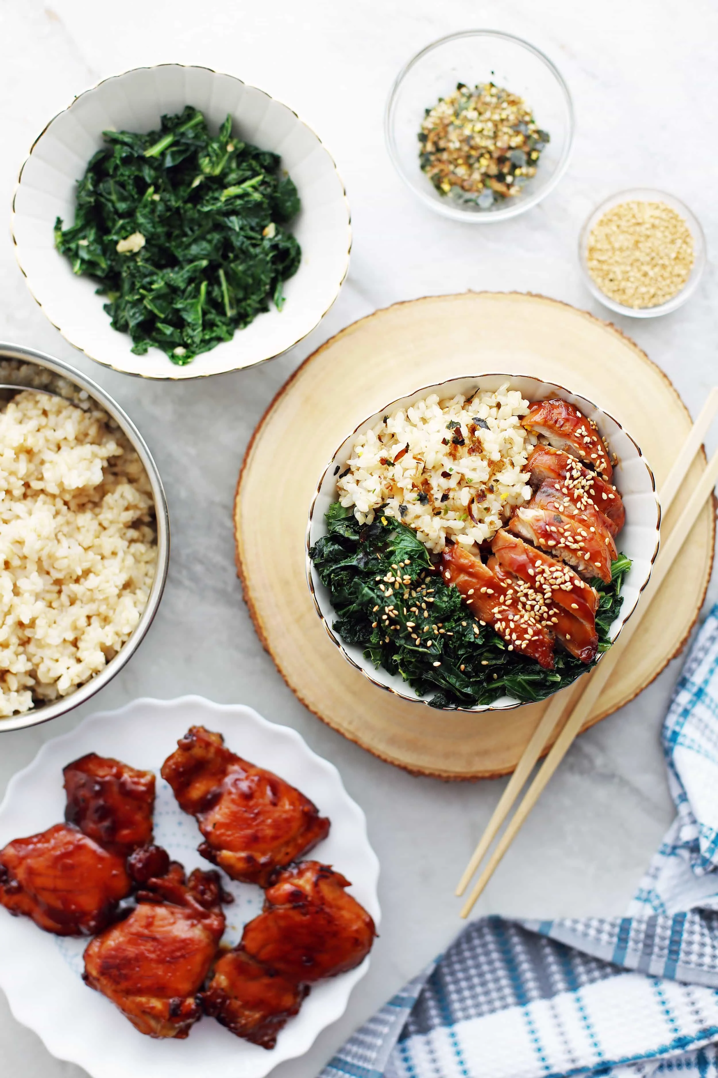 Overhead shot of a bowl of teriyaki chicken with garlicky kale and brown rice, a plate of teriyaki chicken thighs, a bowl of brown rice, and seasonings.