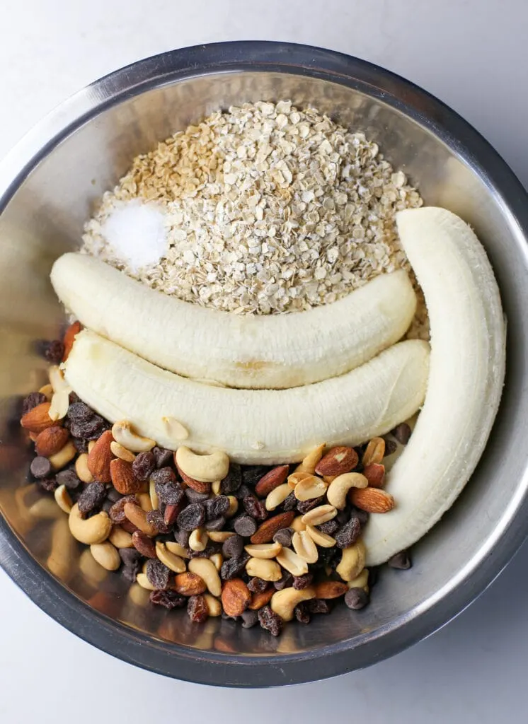 Two peeled bananas, quick oats, trail mix, vanilla extract, and salt in a stainless steel bowl.
