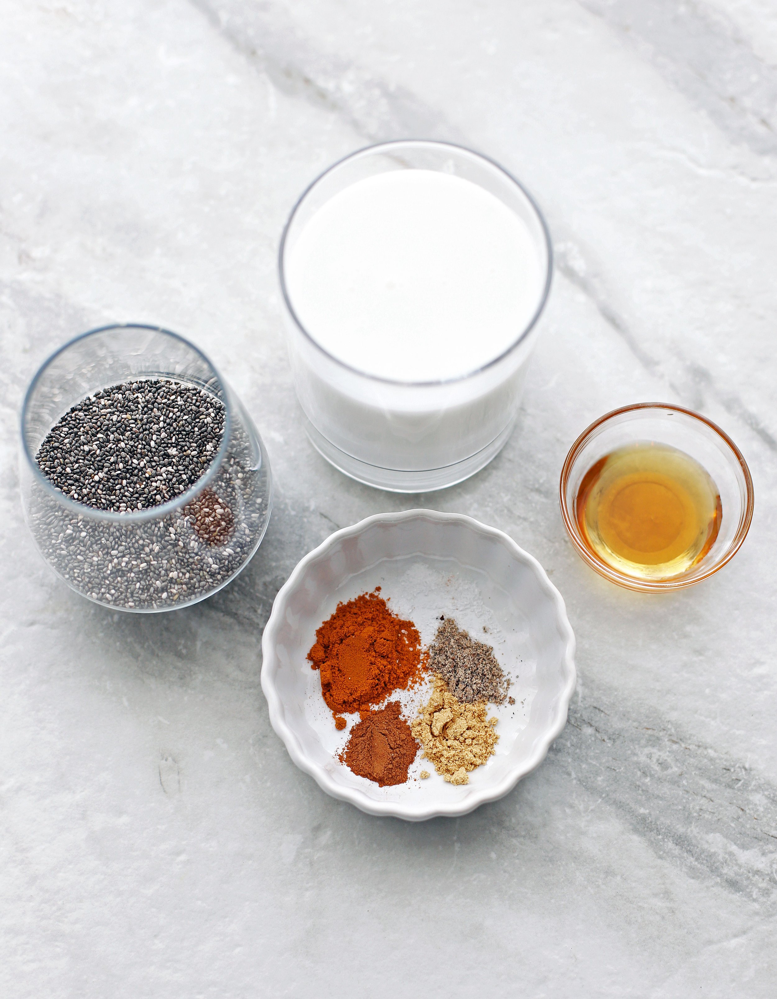 Chia seeds, coconut milk, maple syrup, and spices including turmeric, ginger, cinnamon, and cardamom in glasses and bowls.