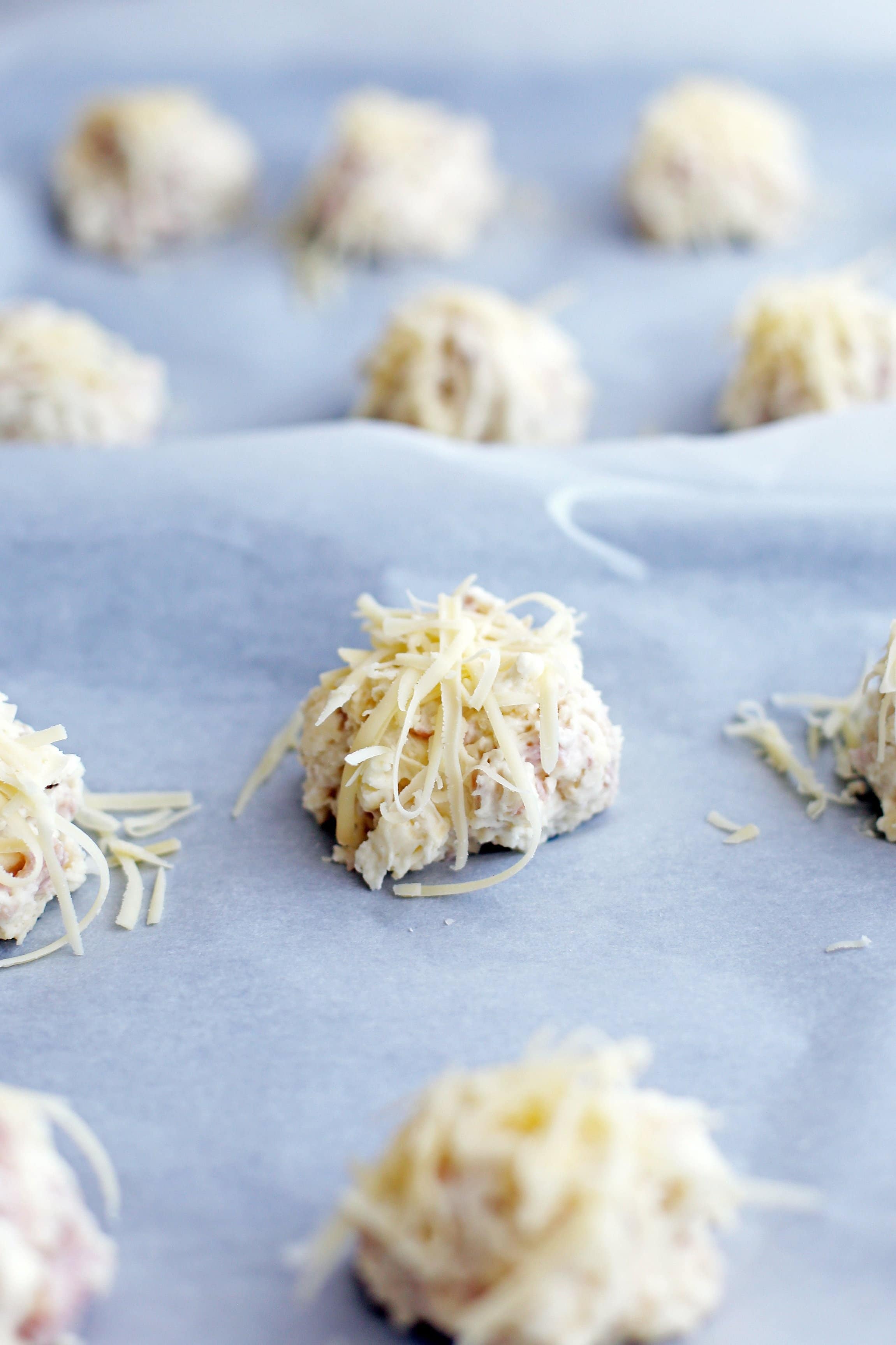 Unbaked ham and cheese drop biscuits topped with shredded gruyere cheese on a parchment paper lined baking sheet.