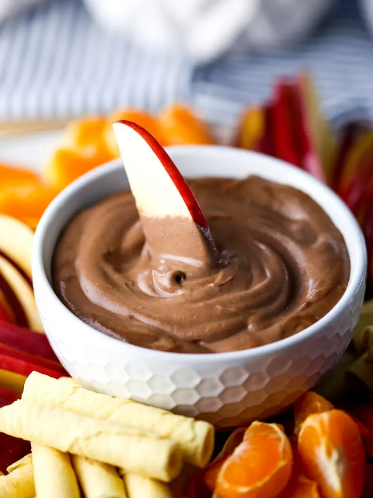 A slice of apple dipped in a white bowl full of creamy cashew chocolate dip.